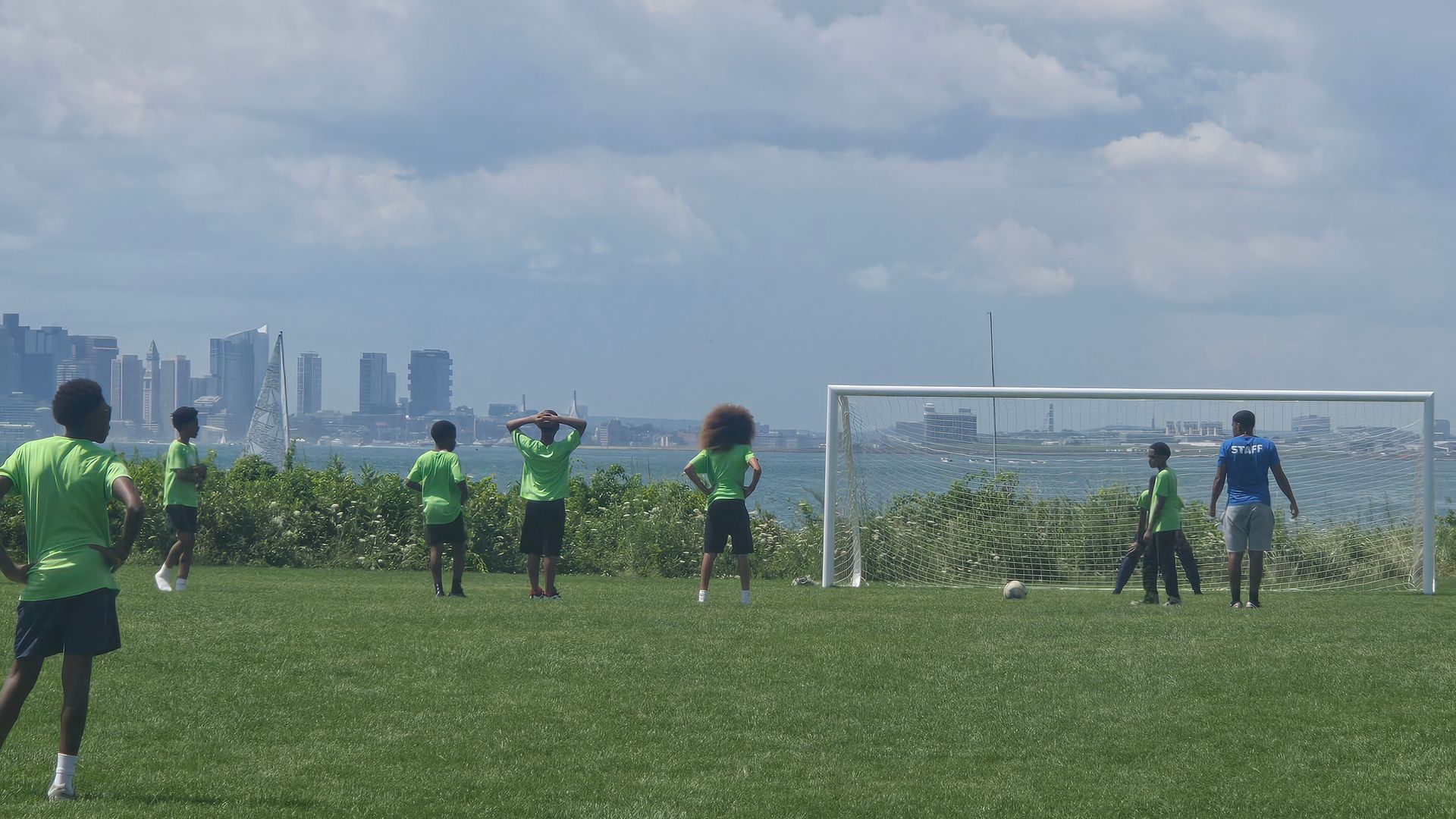 Young players in bright green shirts practice soccer on a grassy coastal field, with a distant city skyline and a goal; a staff member in blue stands by.