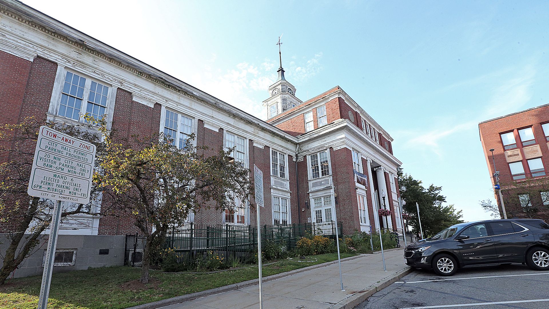Red-brick city hall building with white columns and a tall clock tower under a blue sky, a sidewalk with parking signs, a tree, and a black SUV parked in front.