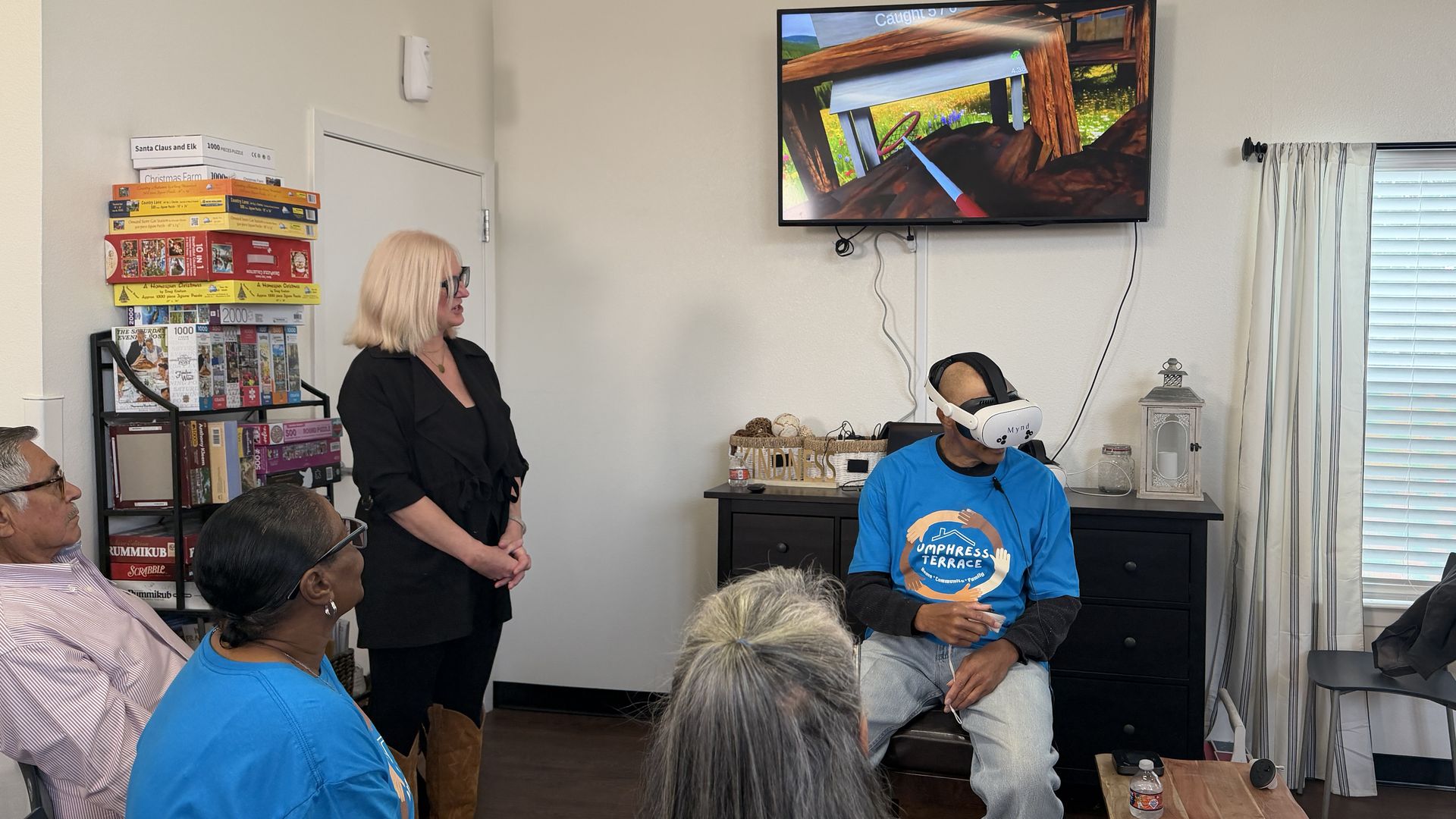 A man wearing a VR headset sits in a room facing a TV screen showing a virtual game; several people watch, including a standing woman with blonde hair and a black outfit.