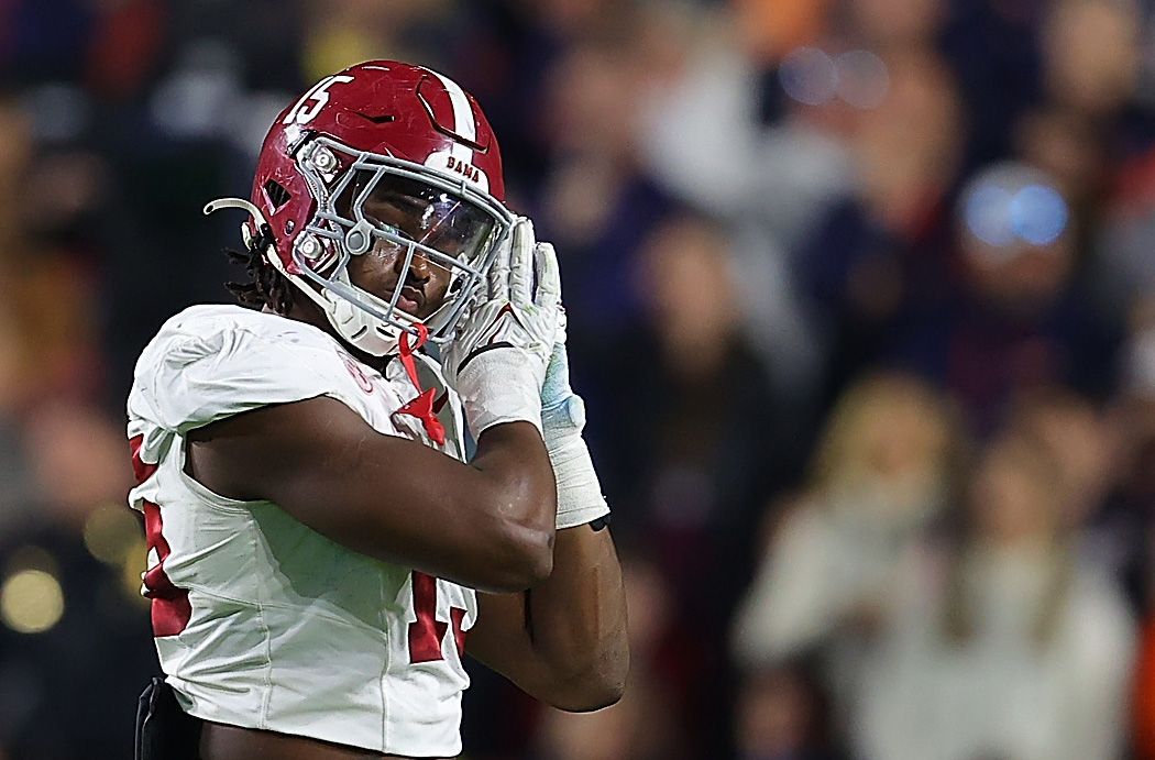 A football player in a white Alabama jersey with a red helmet, number 15, adjusts his facemask while standing on the field; blurred crowd in the background.