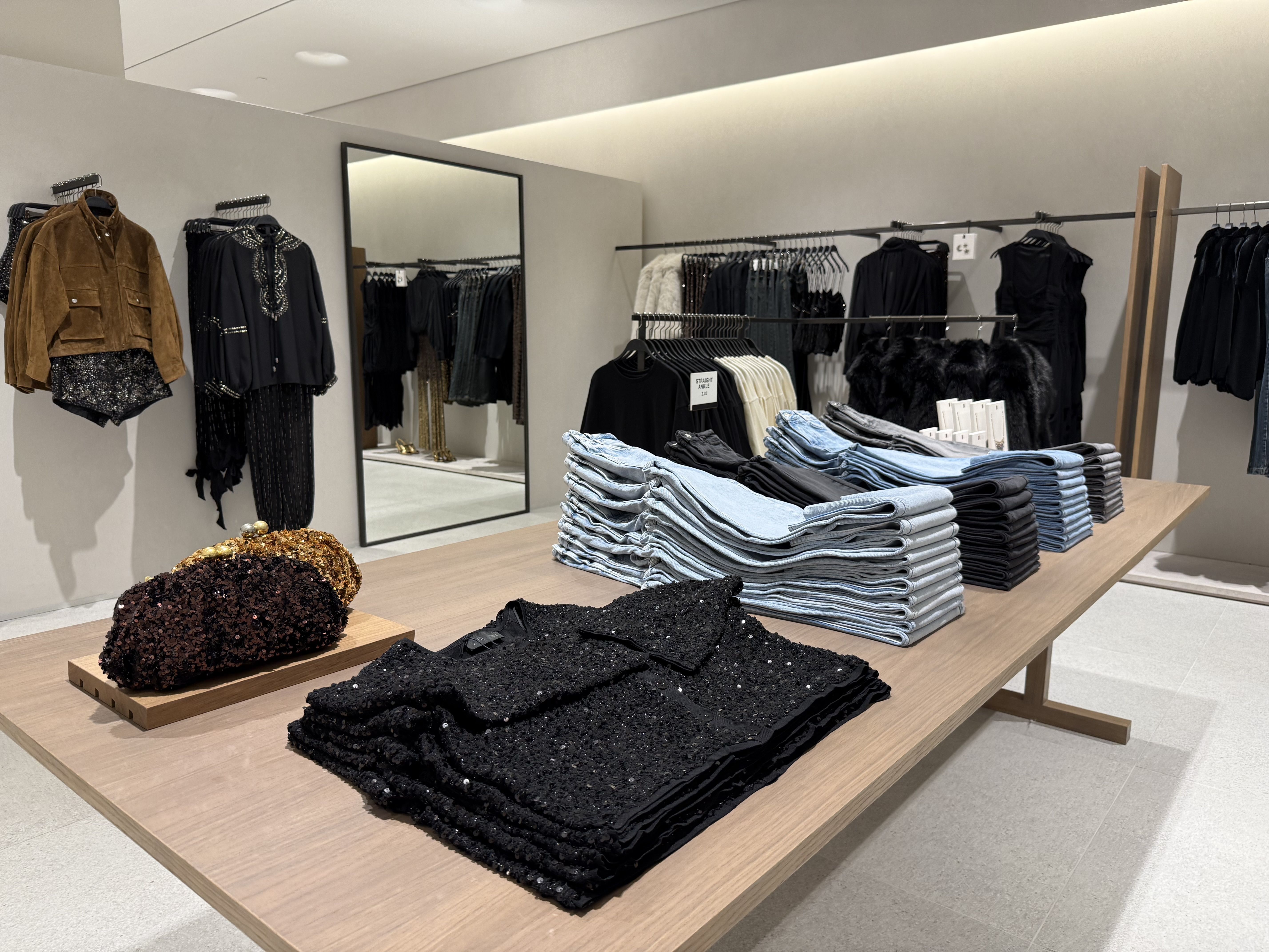 Clothing store display with folded jeans in light blue, black, and gray, black sequined tops, two sequin clutches in black and gold, and hanging brown jacket and black embellished tops against beige walls.