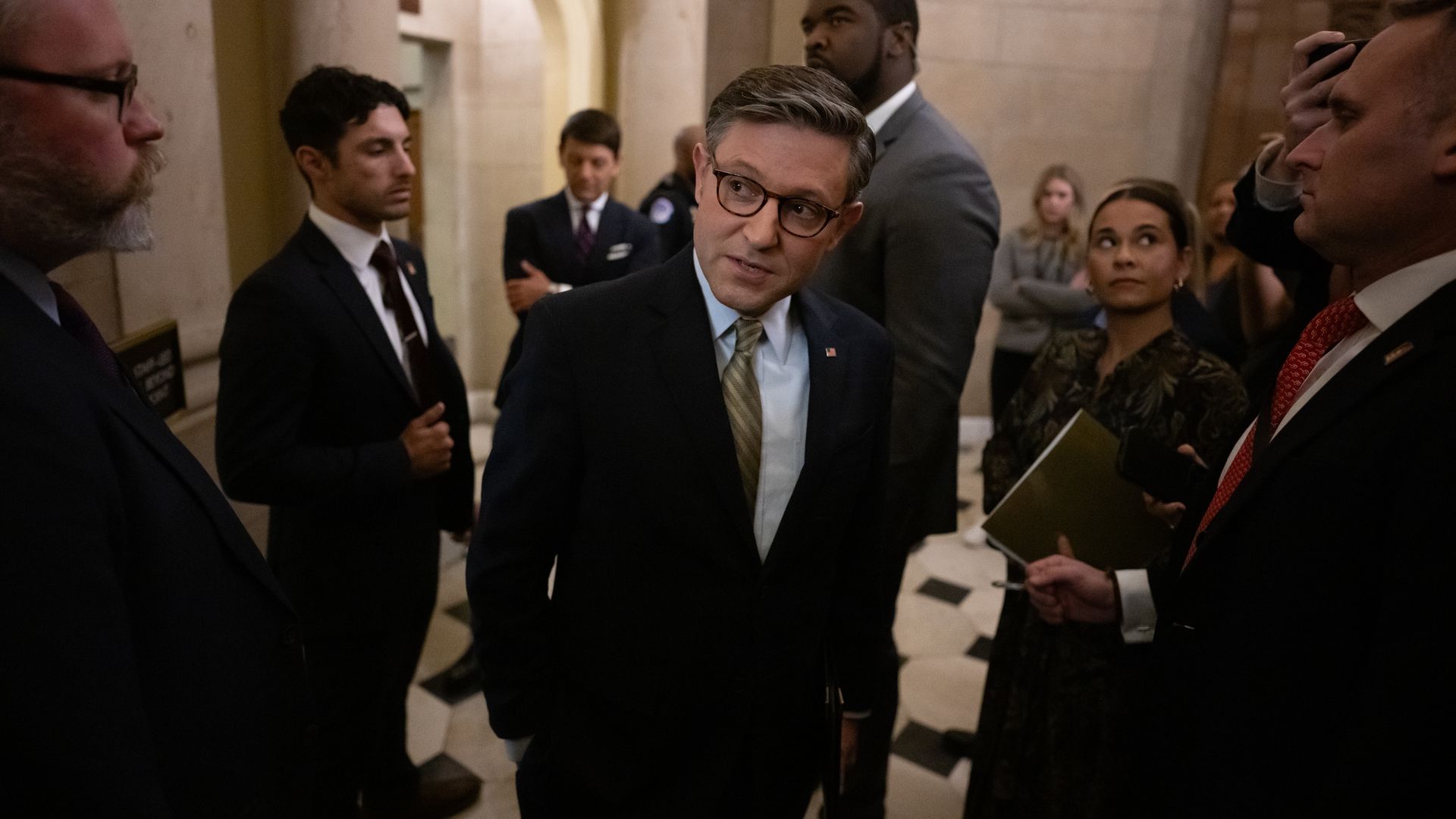 House Speaker Mike Johnson, wearing a gray suit and standing in a sandstone corridor surrounded by staff and security.