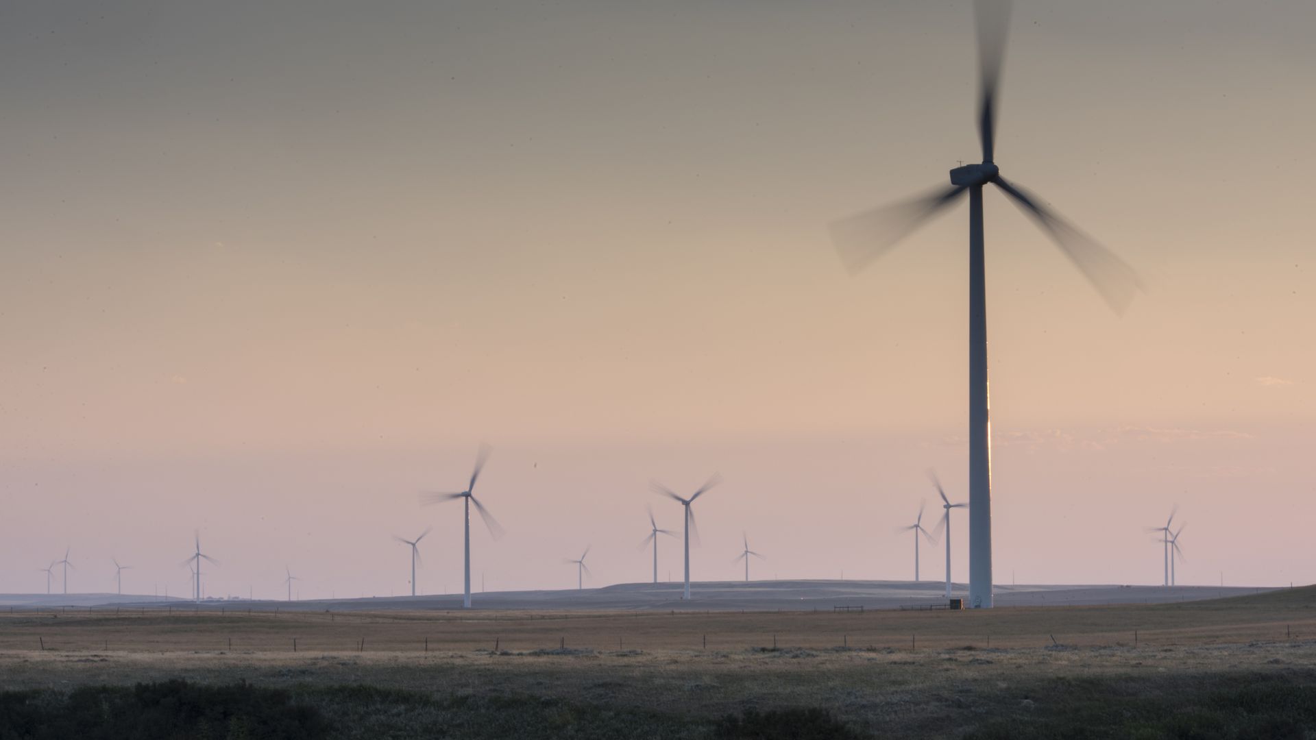 A field of wind turbines spinning in front of a sunset of light pinks and oranges