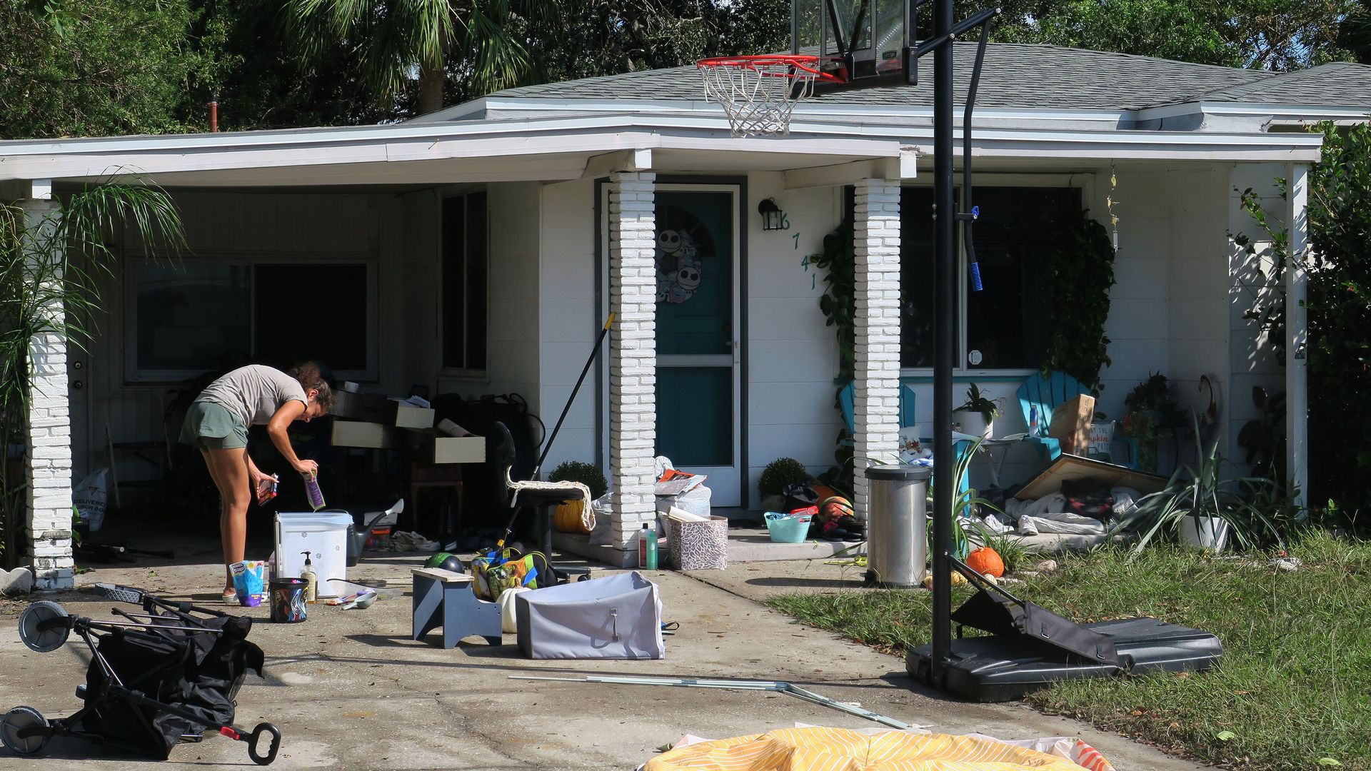 A woman in a gray top and moss-green shorts goes through a bin sitting in her driveway. Her yard is scattered with random items including a laundry hamper and a stroller.