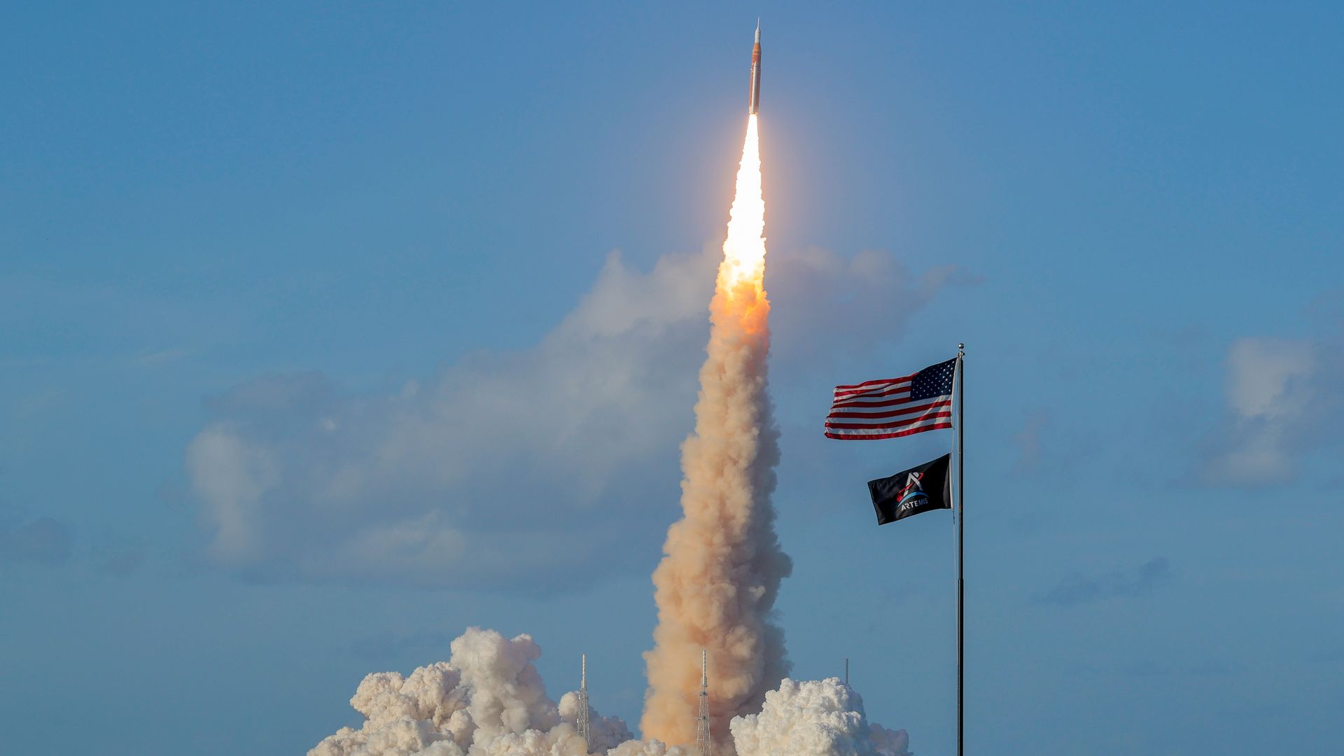 A shuttle launches into the air, with a U.S. flag on the flagpole next to it