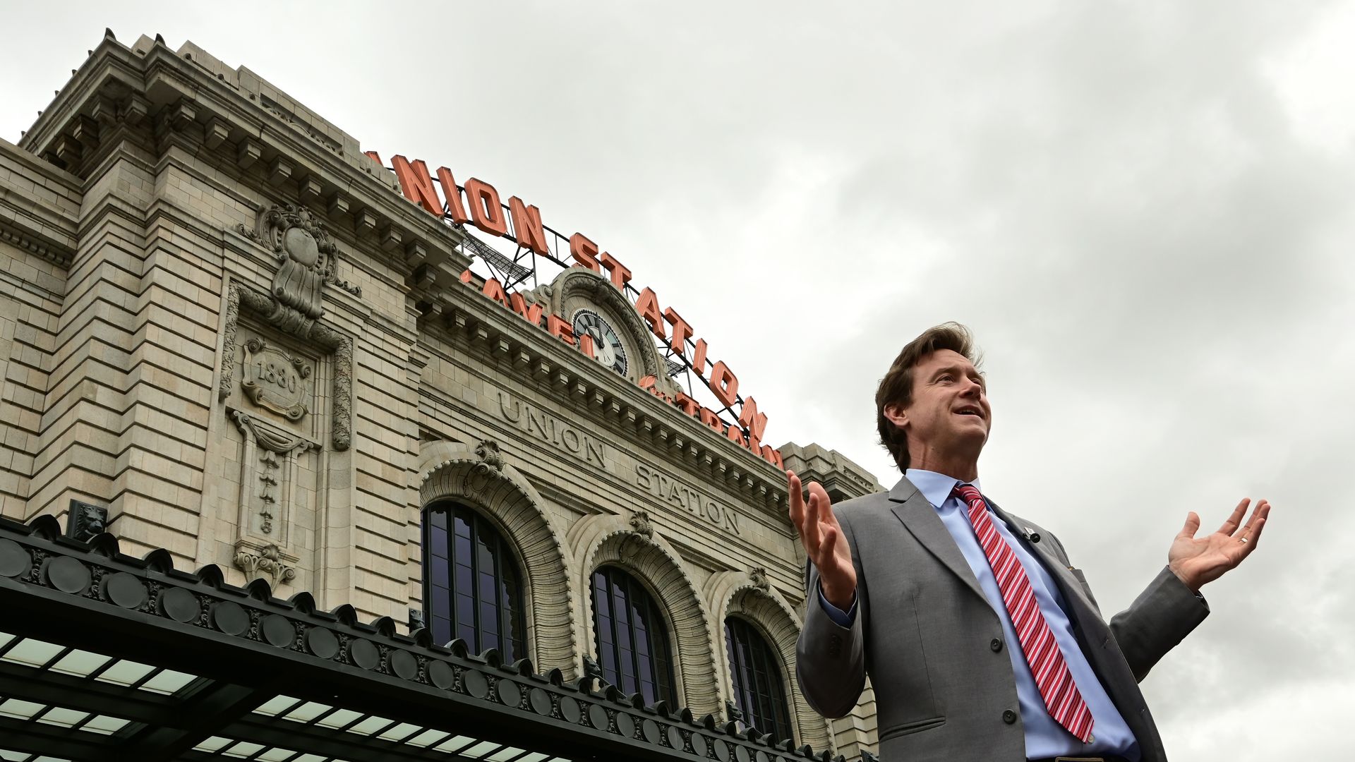 A man in a gray suit, a red-stripped necktie, and a baby blue shirt stands in front of a massive, decorative building with three windows on its facade. The words UNION STATION are engraved in the building. 
