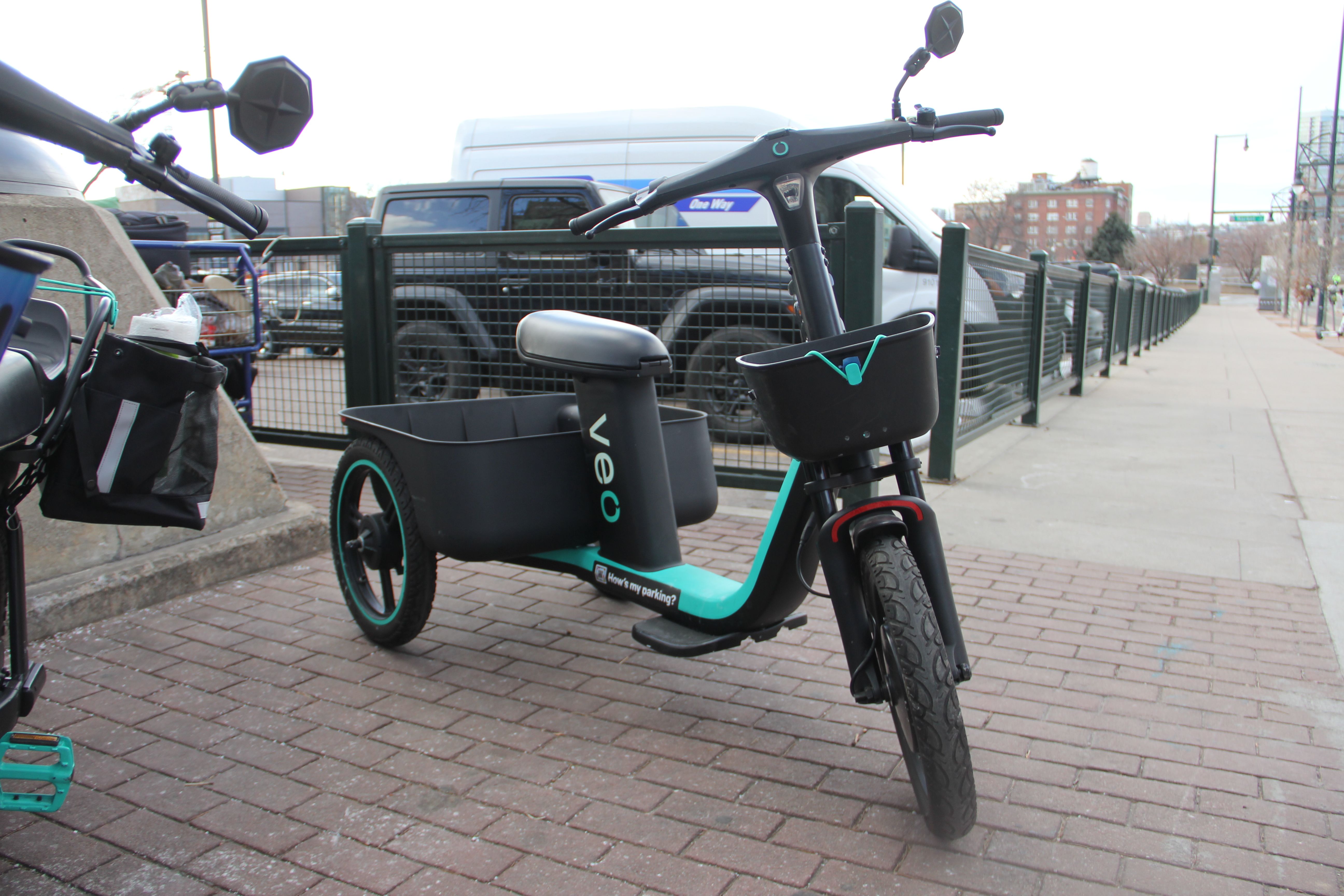 Black and teal Veo electric three-wheeled cargo bike with basket and rear storage compartment parked on a brick sidewalk near a metal fence and street in an urban area.