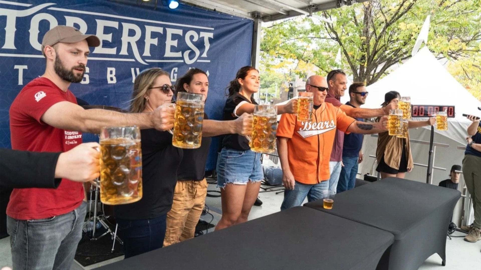 Seven people at Oktoberfest holding large mugs of beer outstretched in front of them, standing side by side on a stage with a banner behind them.