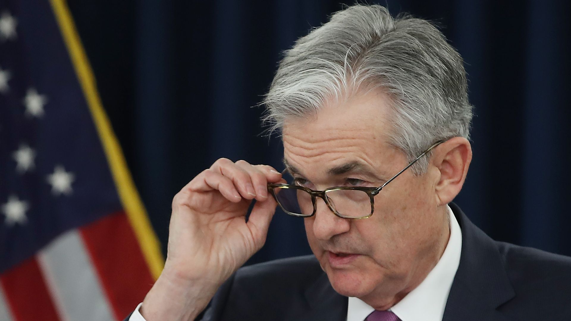 In this image, Jerome Powell looks over his glasses while speaking and standing in a suit.
