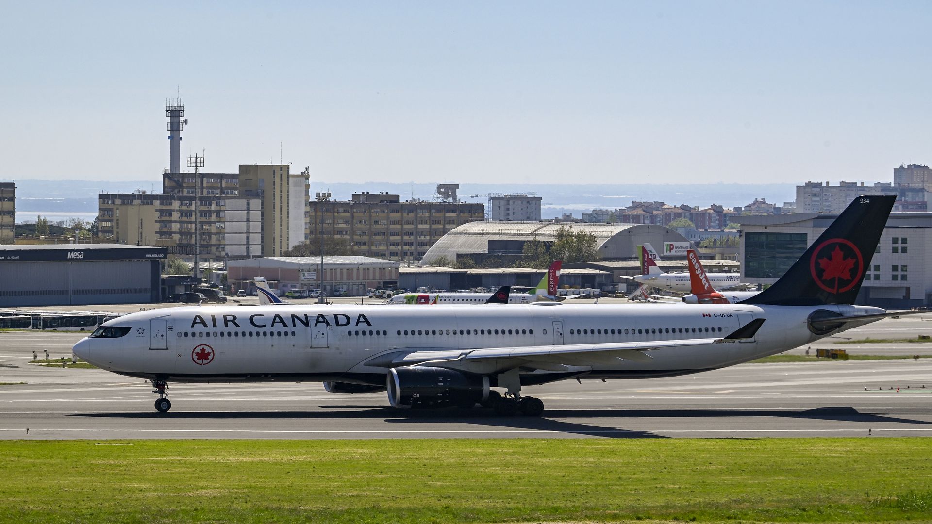 Air Canada passenger jet on the taxiway with white fuselage and a red maple leaf on the tail; city buildings and a control tower rise in the background under a clear blue sky.