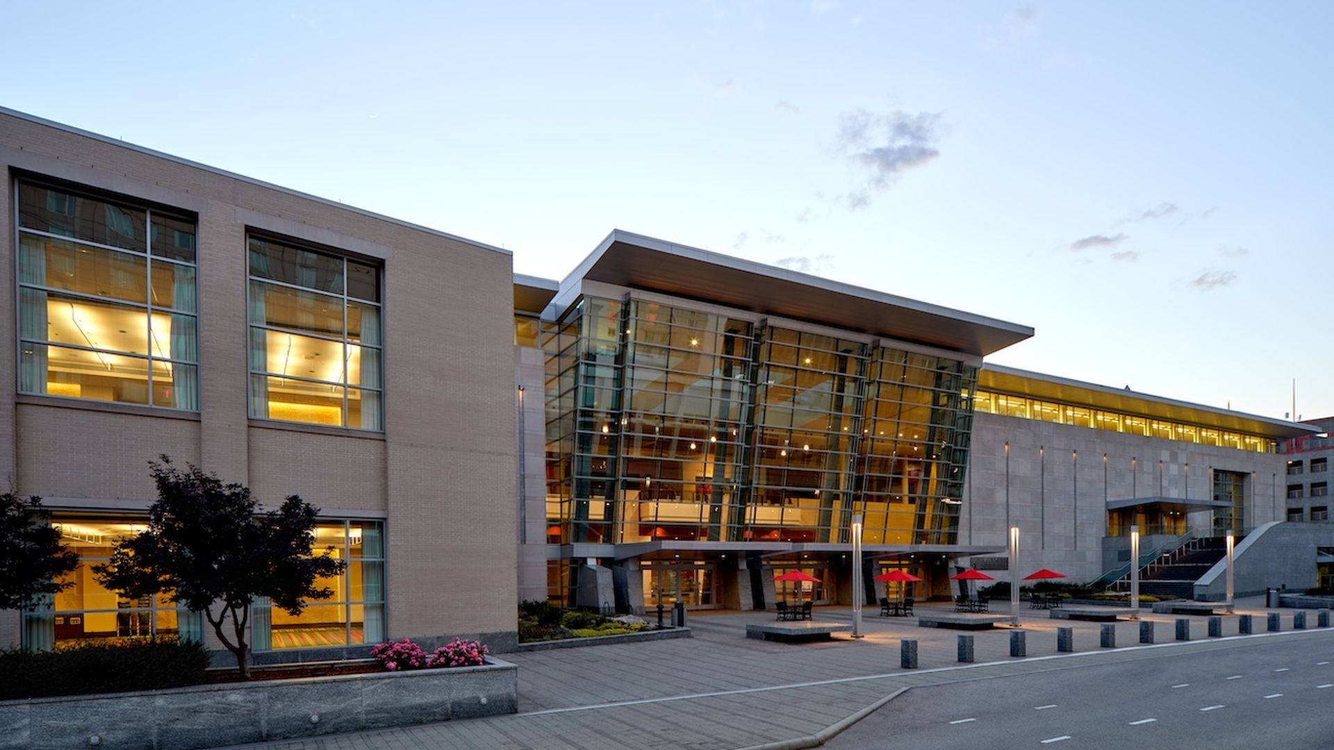 Modern campus building featuring a glass, angular atrium, beige brick walls, and large windows. Outdoor seating with red umbrellas along a paved plaza, with trees and pink flowers nearby.