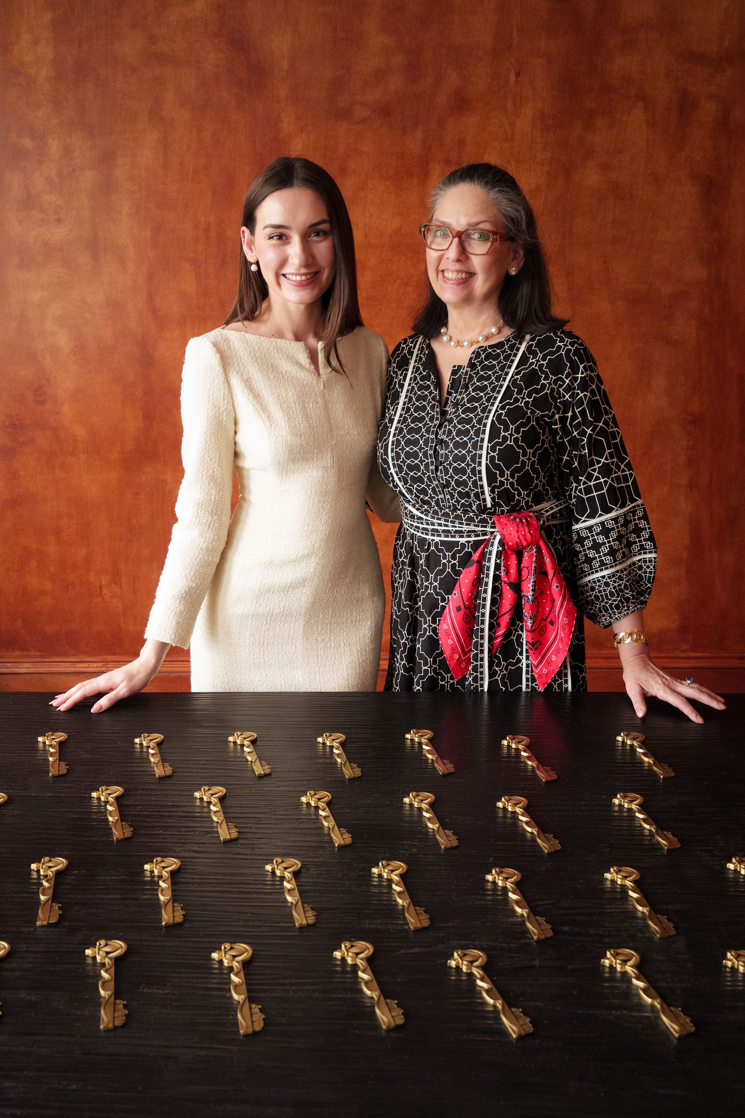 Two smiling women stand behind a dark table covered with gold keys arranged in rows; warm brown backdrop. Left: cream dress, right: black patterned dress with a red sash.