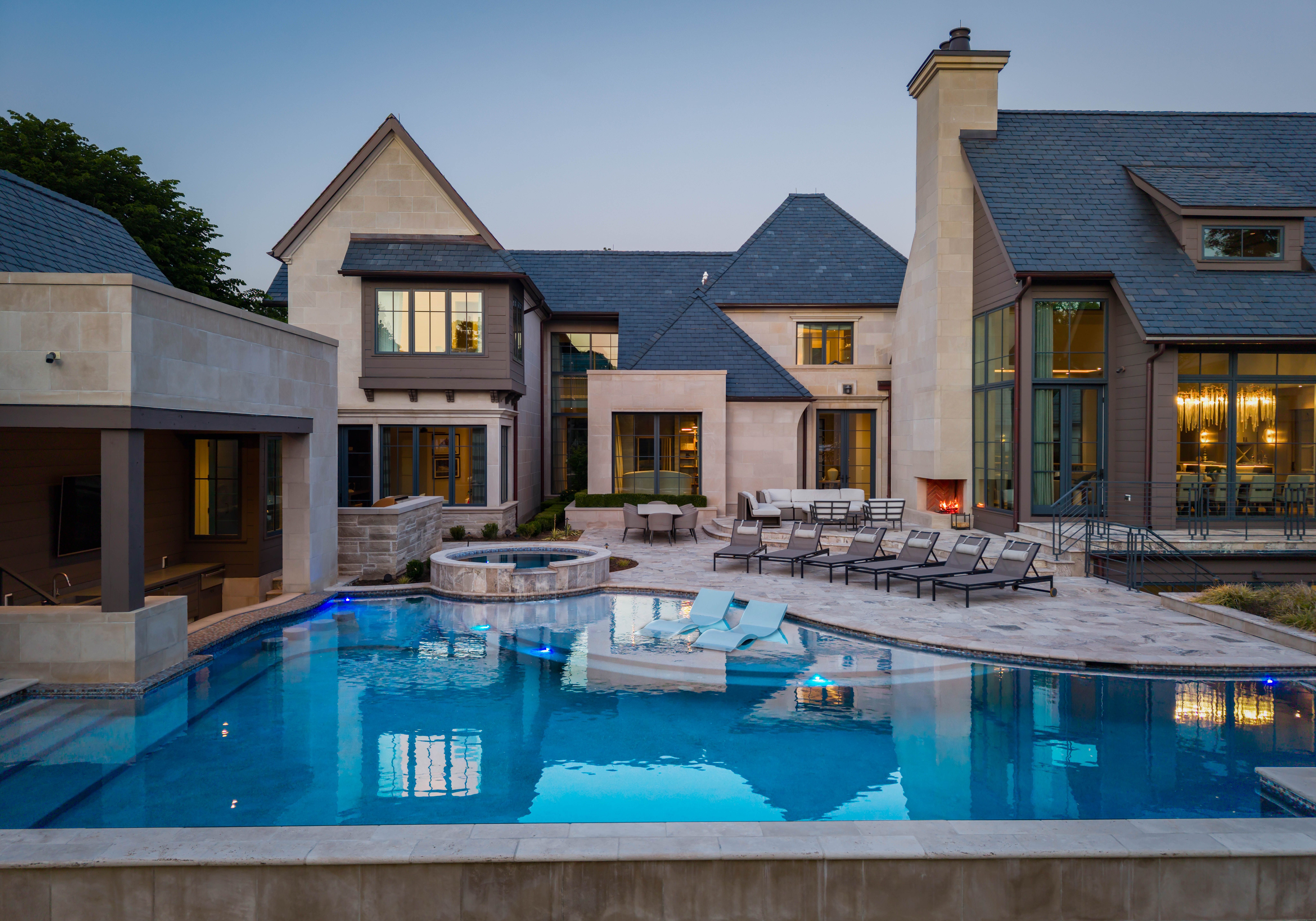 Luxury modern house with blue slate roofs and beige stone walls. Central courtyard features a rectangular pool, circular hot tub, glass doors, and numerous lounge chairs and outdoor seating.