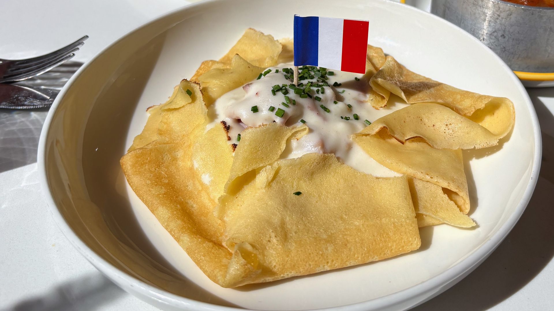 A folded crepe with white cream sauce and chopped chives, topped with a small French flag, served on a white plate in natural light next to a fork and a bowl of tomato-based stew.