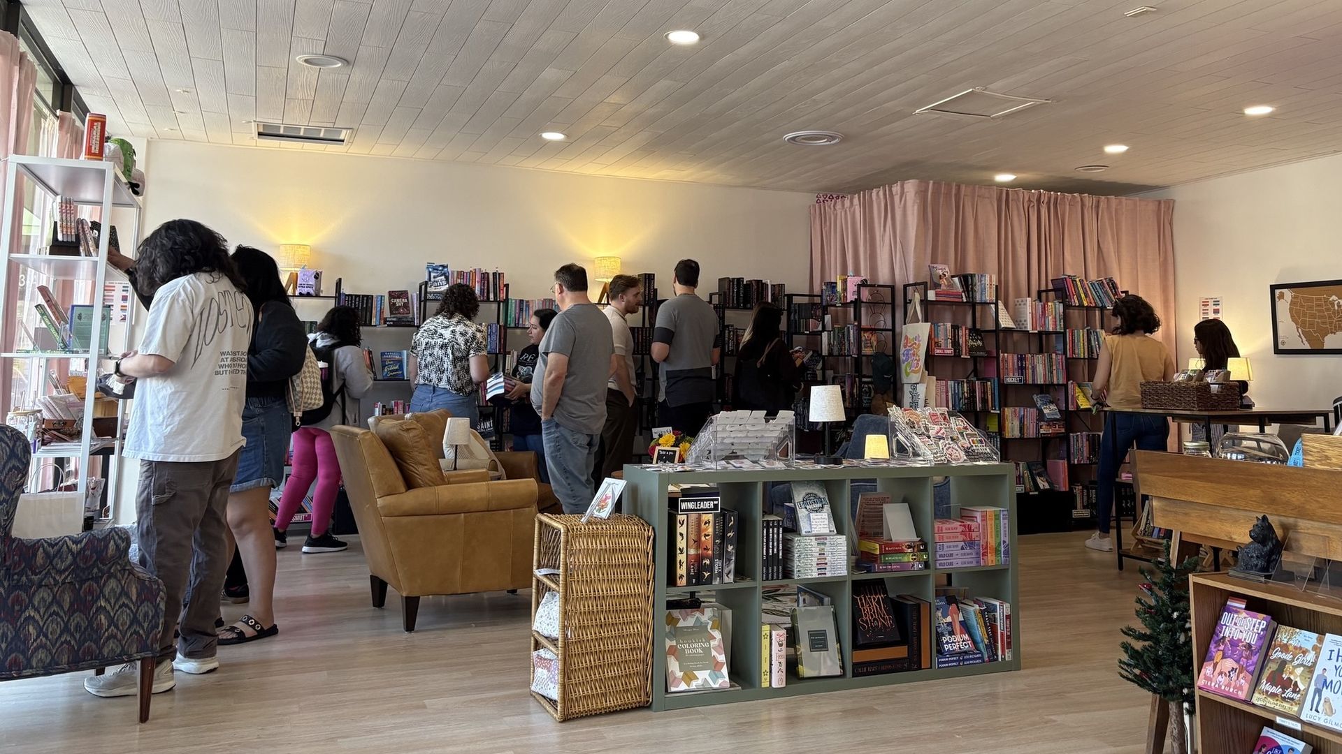 Interior of a bookstore with people browsing books on black metal shelves, a light brown armchair, wooden floor, softly lit lamps, and a small green bookshelf displaying various books.