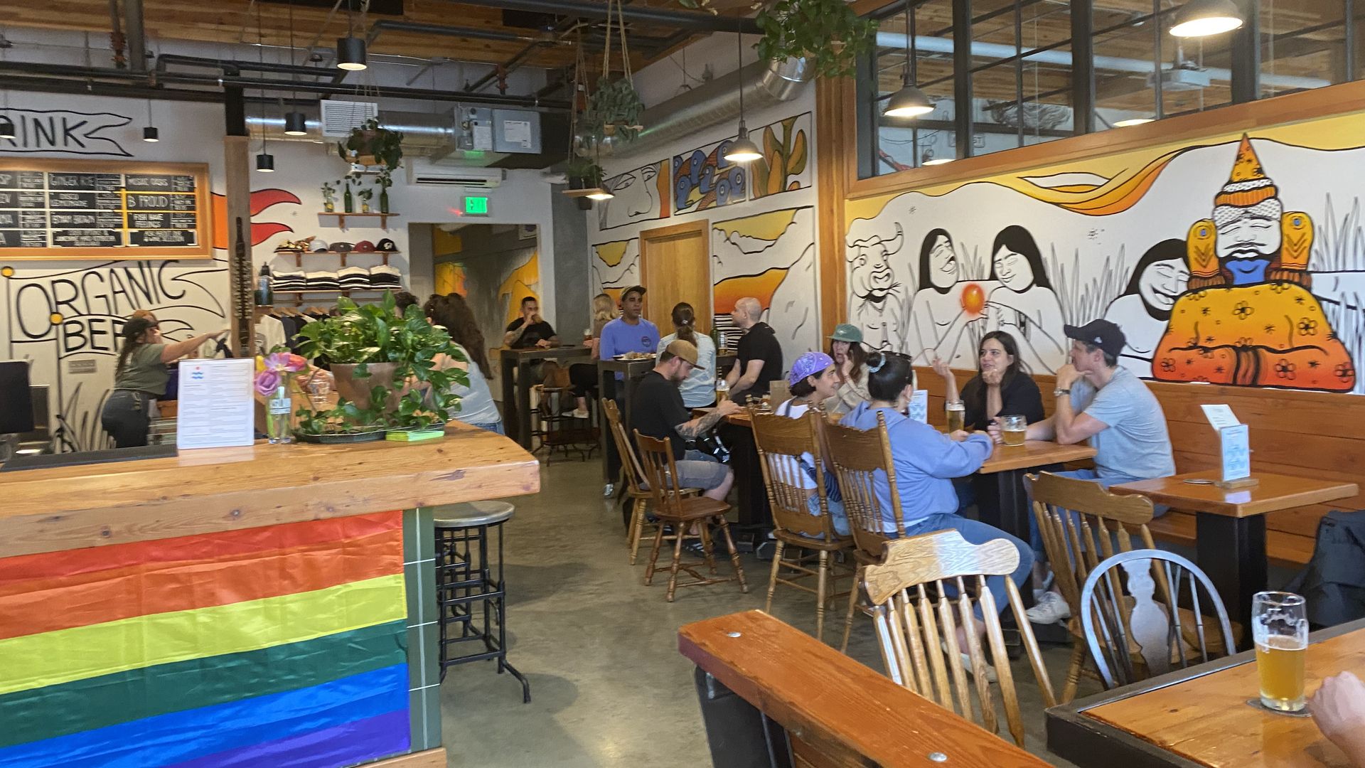 A view of the interior of a tap room with murals on the walls and people at small tables, and a pride flag at the end of the bar.