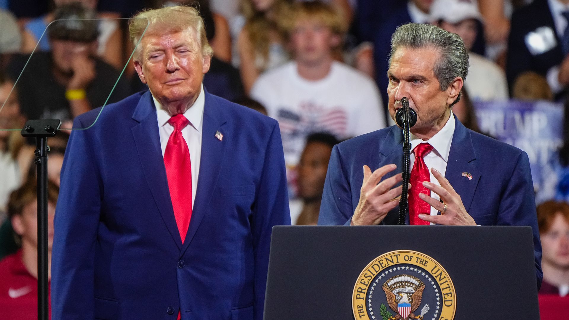 A man in a dark blue suit, white shirt and red tie speaks at a microphone and podium adorned with the President of the United States seal. Another man stands to his right and wears a dark blue suit, red tie and white shirt.