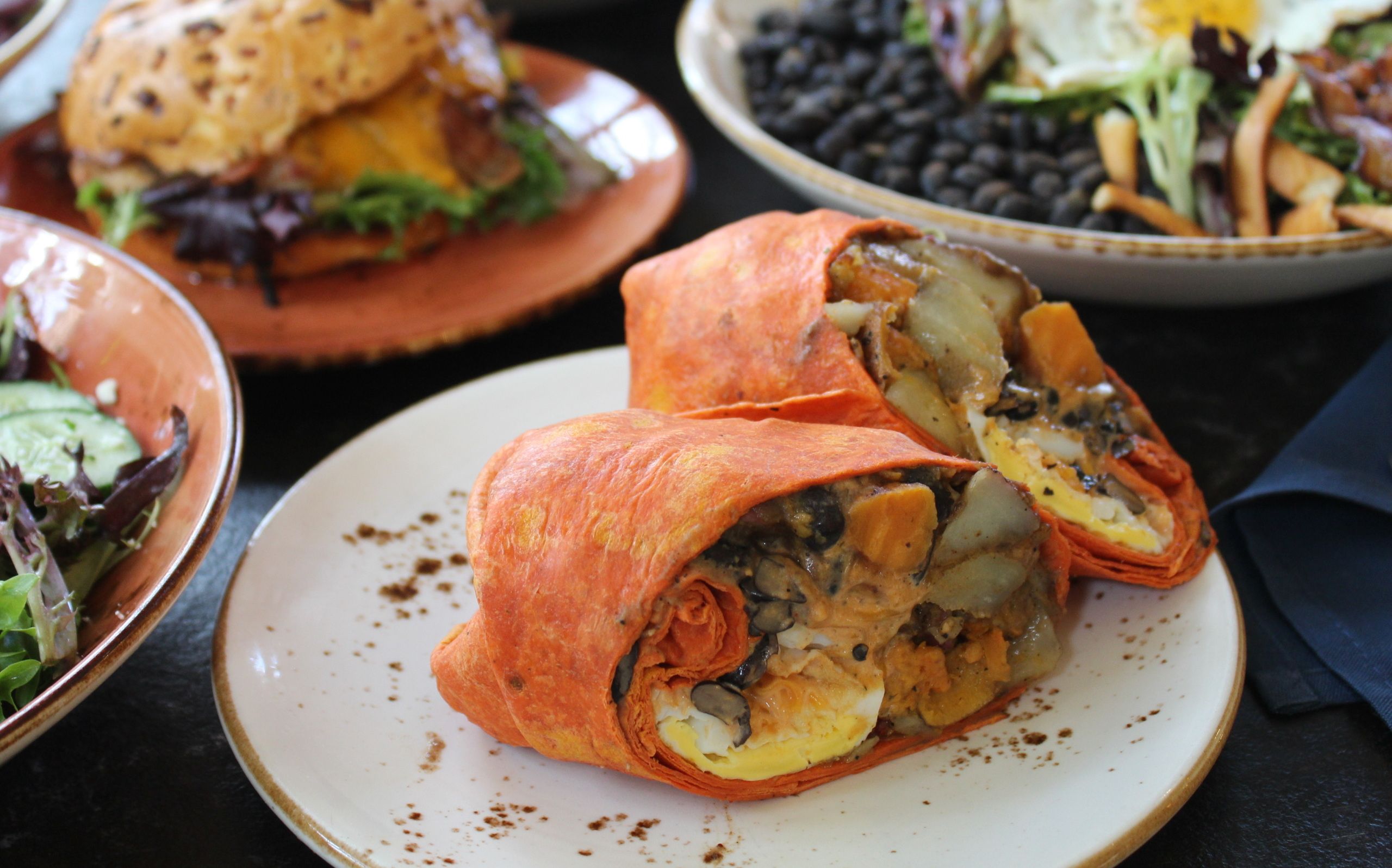 Cut orange wrap filled with egg, black beans, potatoes, and vegetables on a white plate, with a sandwich and salad in the background on a dark table.
