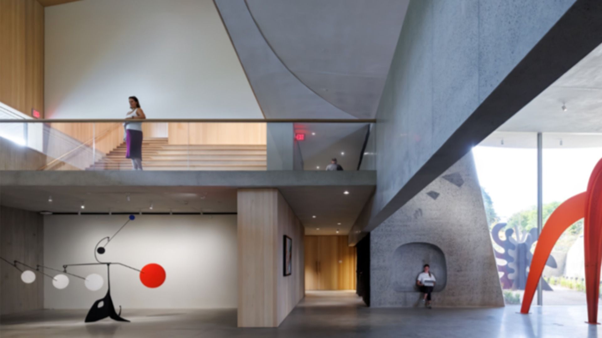 The inside of the building at Calder Gardens, which has a modern interior with concrete and wood elements, a large red and black mobile sculpture, people on upper and lower levels, and a bright space with large windows revealing outdoor sculptures.