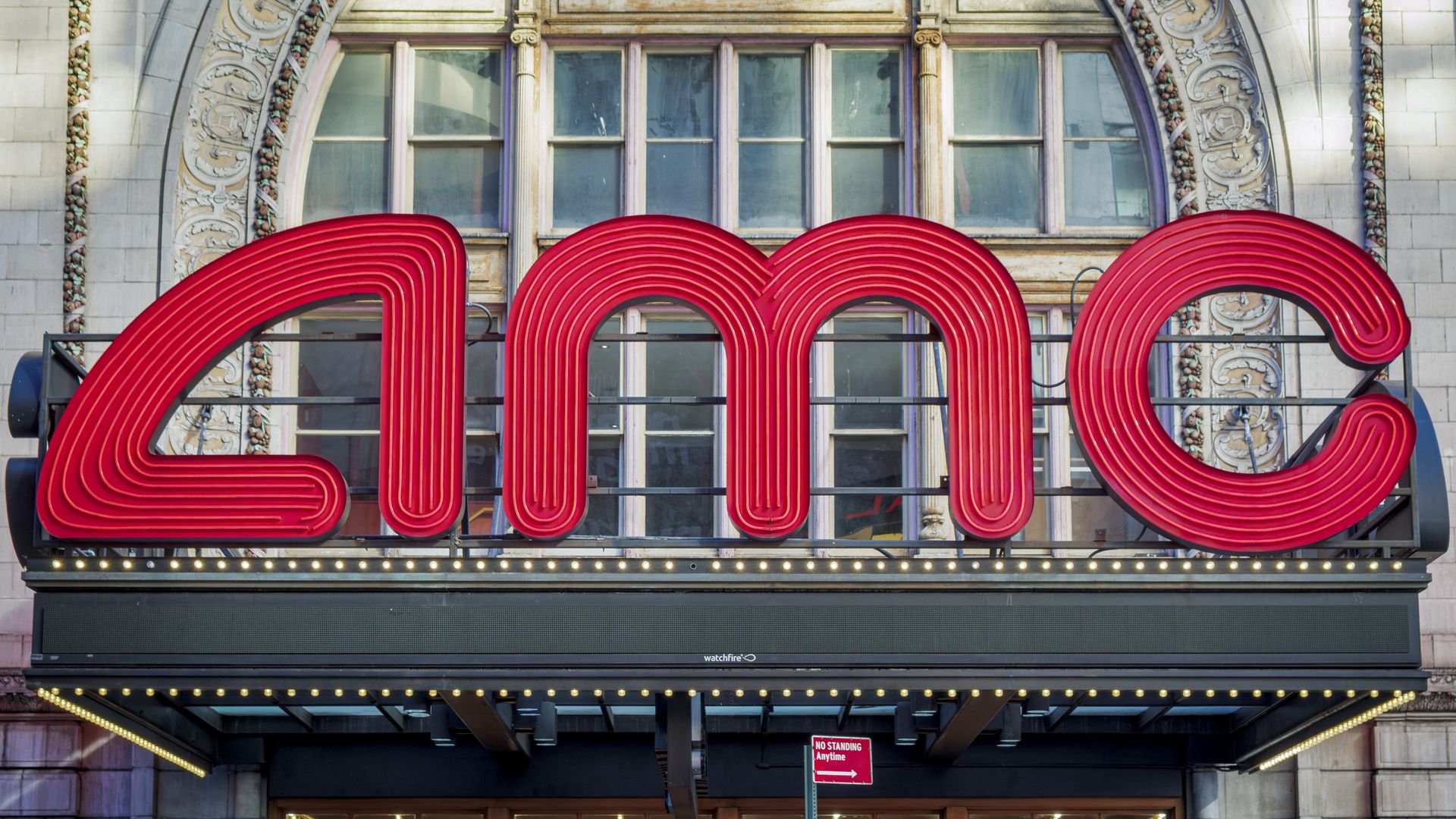 A large red neon sign with multi-layered tubular letters sits above a storefront, backed by an ornate stone building with arched windows; a bulb-lit marquee runs along the bottom.