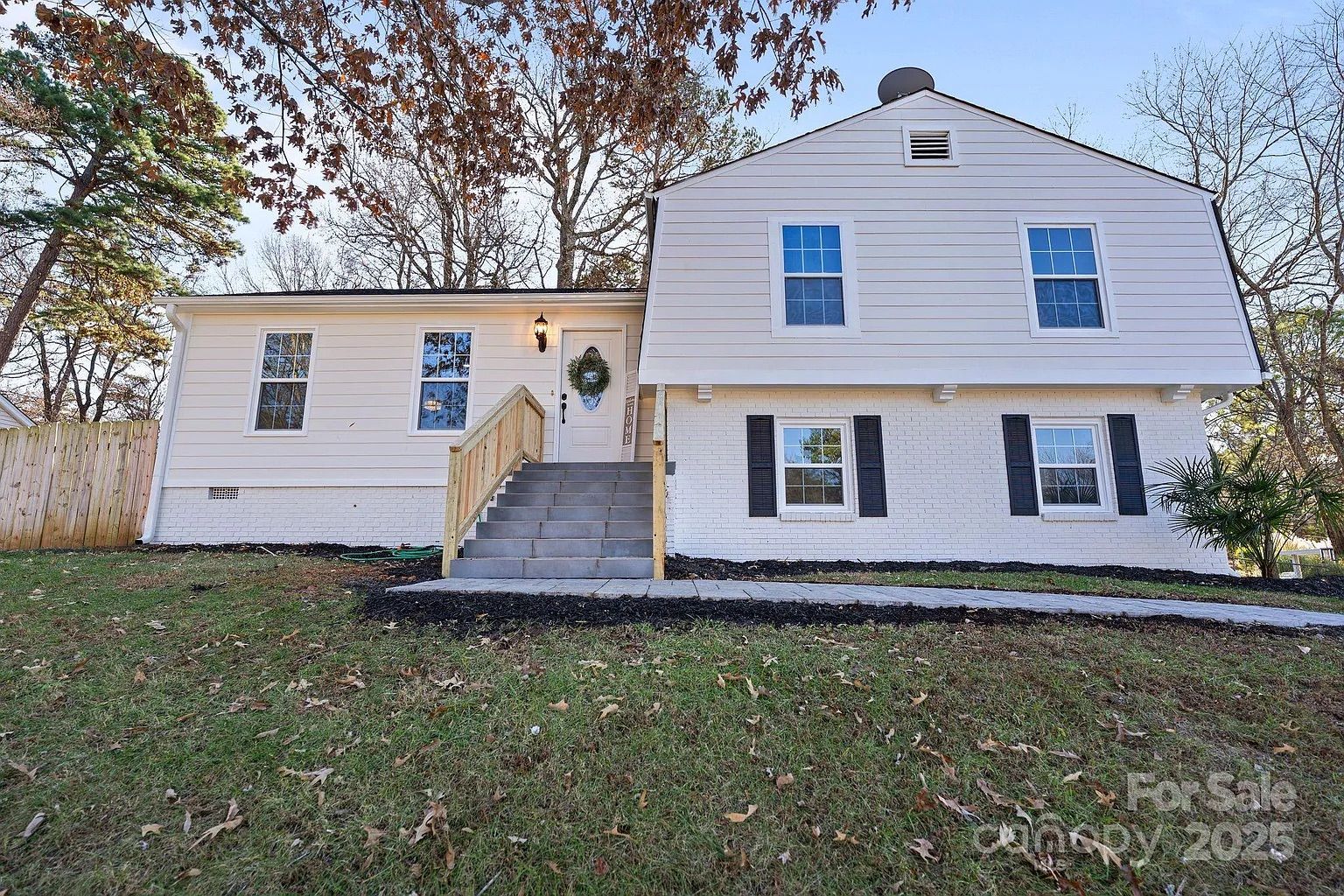 White split-level house with six windows, a front door with a green wreath, wooden railing stairs, lawn with fallen leaves, trees in background, and a "For Sale" sign.