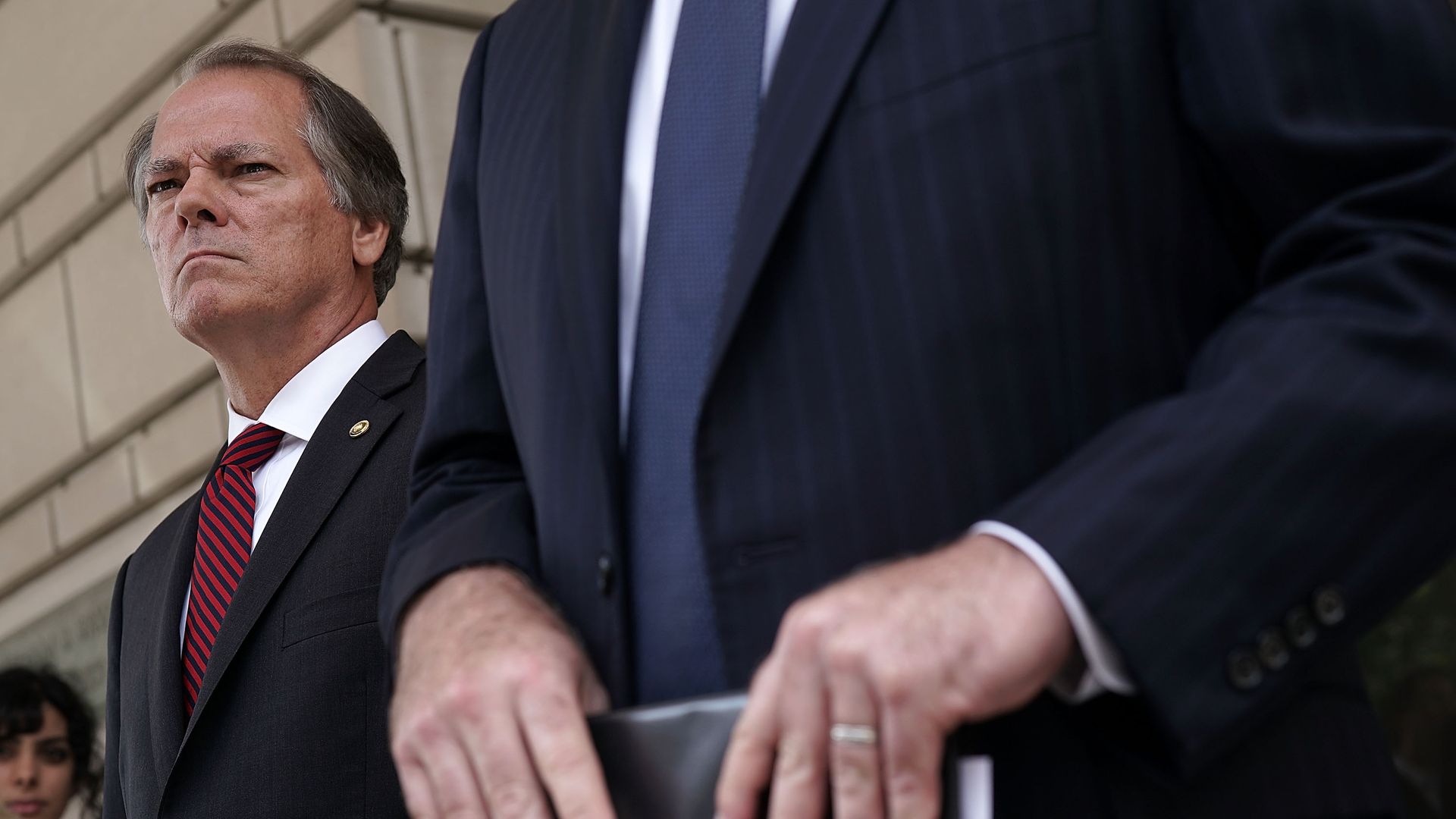 James Wolfe looks angrily into the crowd with the hands of his lawyer on a folder in the foreground.