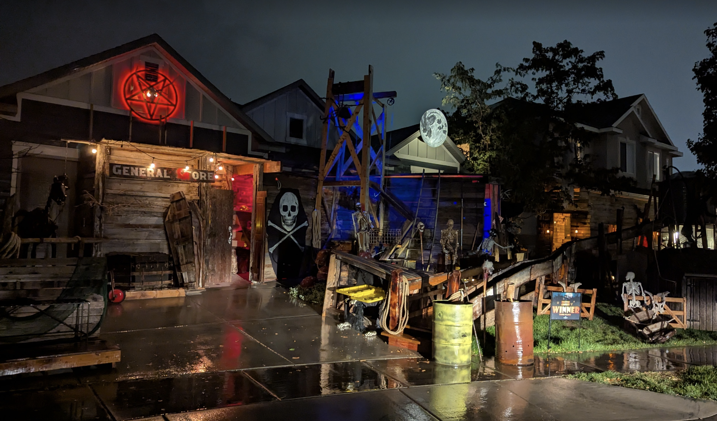 Nighttime Halloween yard with a weathered storefront sign reading GENERAL STORE, a red neon pentagram, skeletons and skulls, wooden props, and a large illuminated moon against a blue-lit background.