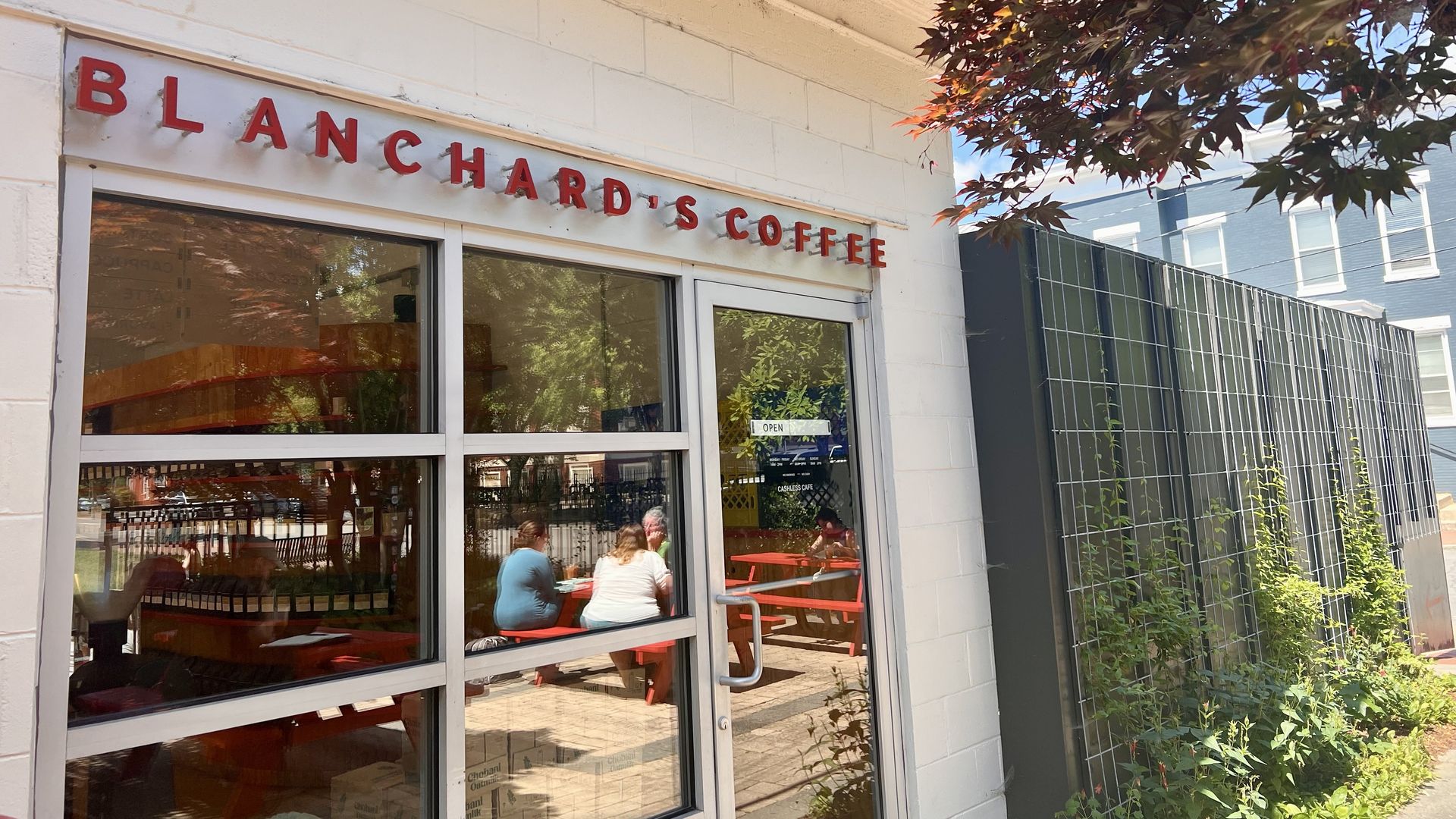 Front of a cafe with a white brick facade and a red Blanchard's Coffee sign. Glass doors reveal red tables and customers inside; a tree with red leaves overhangs the entrance; wall light above.