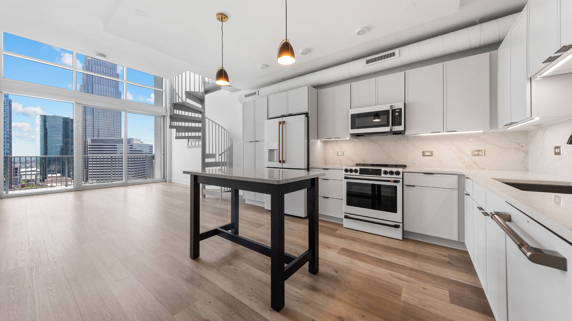 Bright modern kitchen with white cabinets, stainless steel appliances, marble backsplash, and a black island. A spiral staircase and large windows overlook a city skyline.