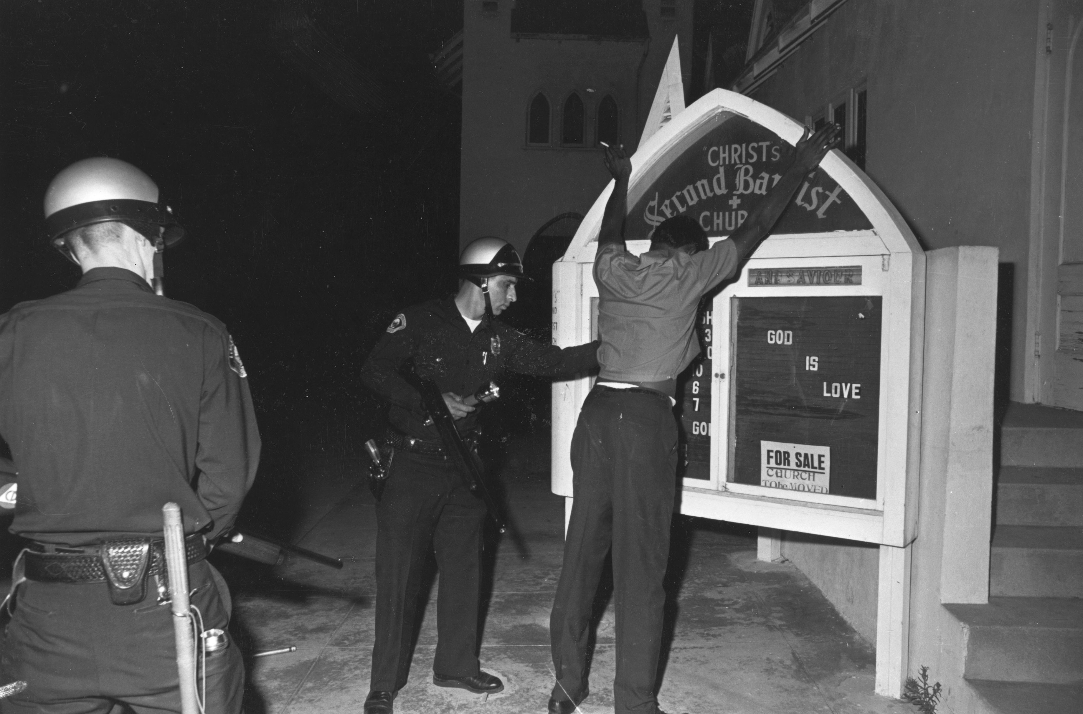 Two police officers frisk a Black man at night in front of a church marquee that says "God is Love" and "For Sale Church to be Moved." The man stands with arms raised.