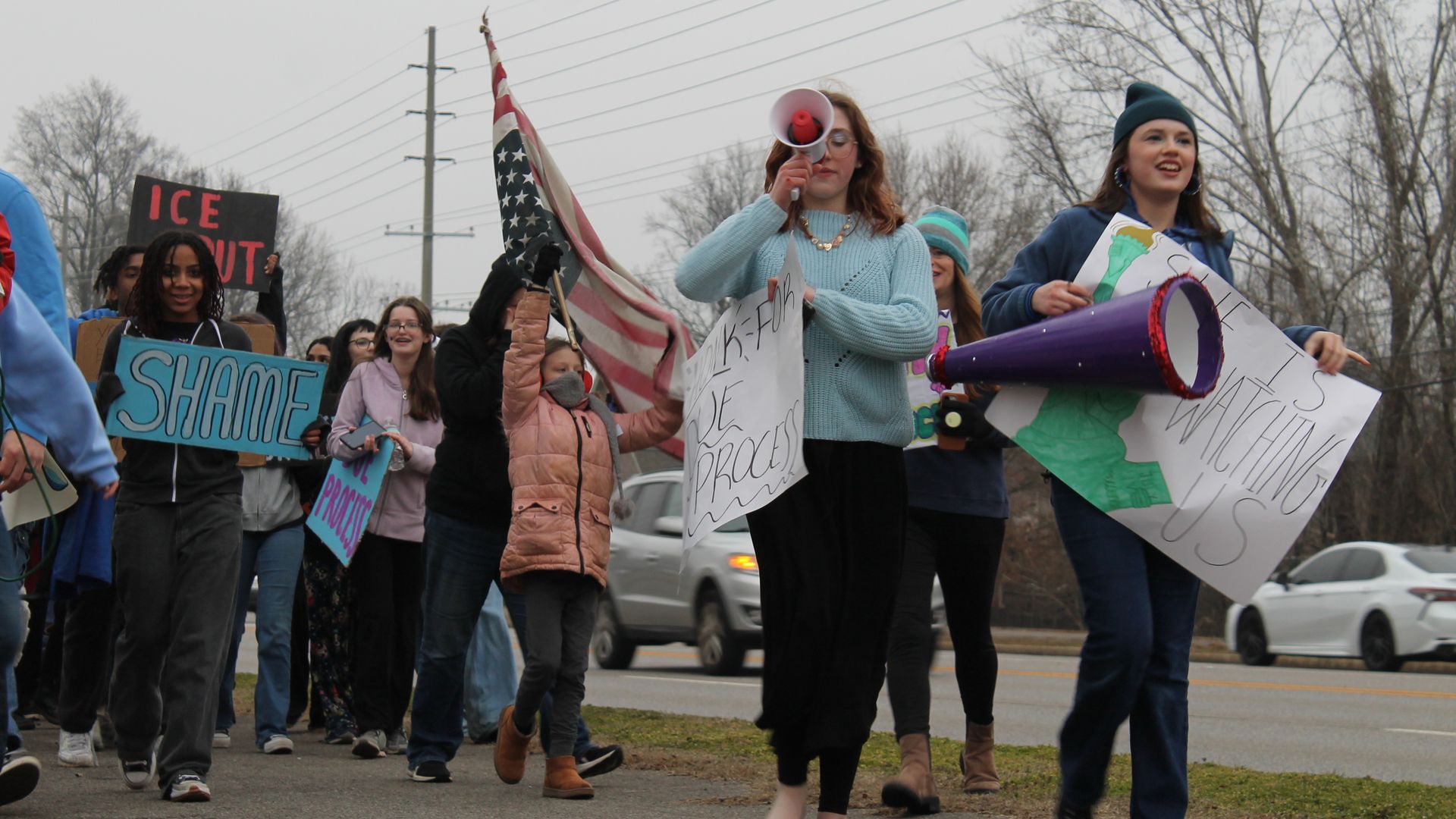 Group of diverse protesters walking on a roadside holding signs and an American flag, one with a megaphone, on an overcast day with bare trees and passing cars in background.