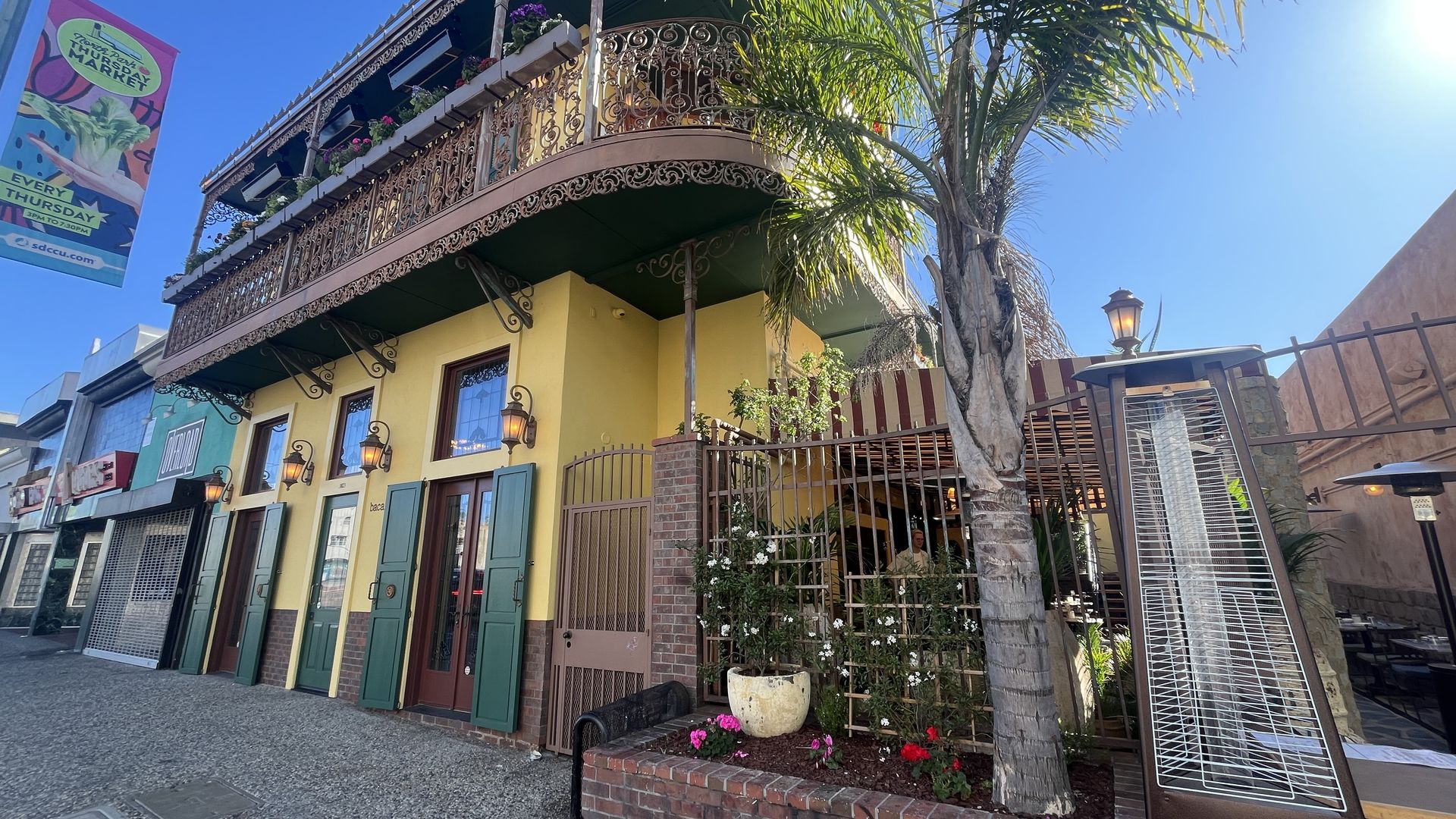 Sunlit street with a yellow two-story building, iron balcony, green shutters, and lanterns. A palm tree beside a brick fence with potted flowers and a striped awning over outdoor patio.