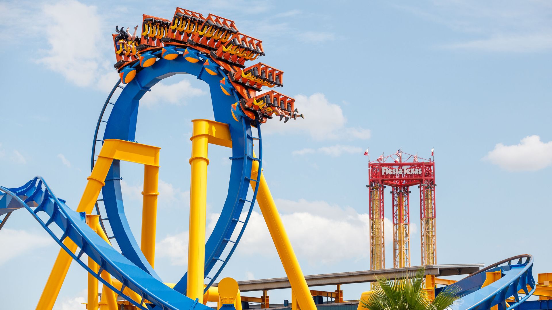 A blue and yellow roller coaster against a light blue sky with clouds at Six Flags Fiesta Texas is seen largely empty in June 2020.