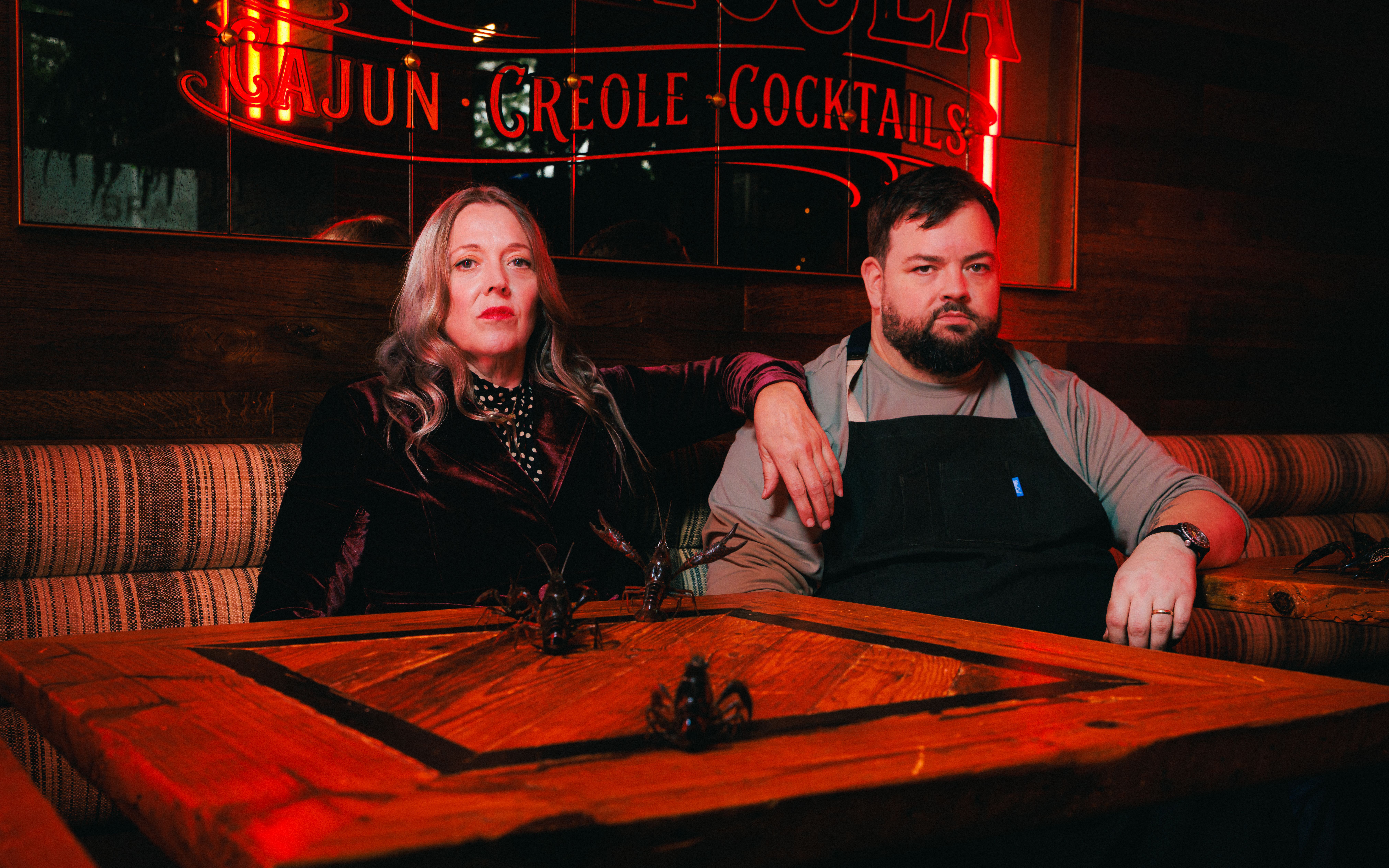 Two people sit on a striped booth in a dim bar under a red neon sign reading "CAJUN CREOLE COCKTAILS." A blonde woman in velvet and a bearded man in a gray shirt with an apron look at the camera.