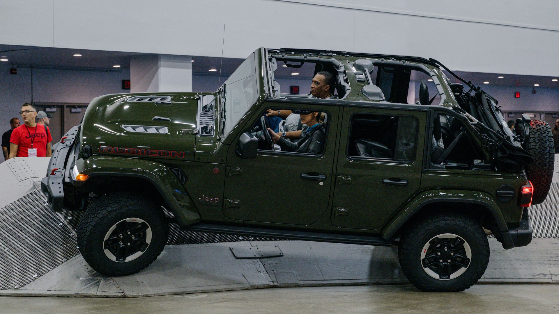 A dark green Jeep SUV with doors off and roof removed on an indoor display. A man sits in the driver’s seat and a woman in the front passenger seat, with large off-road tires visible.