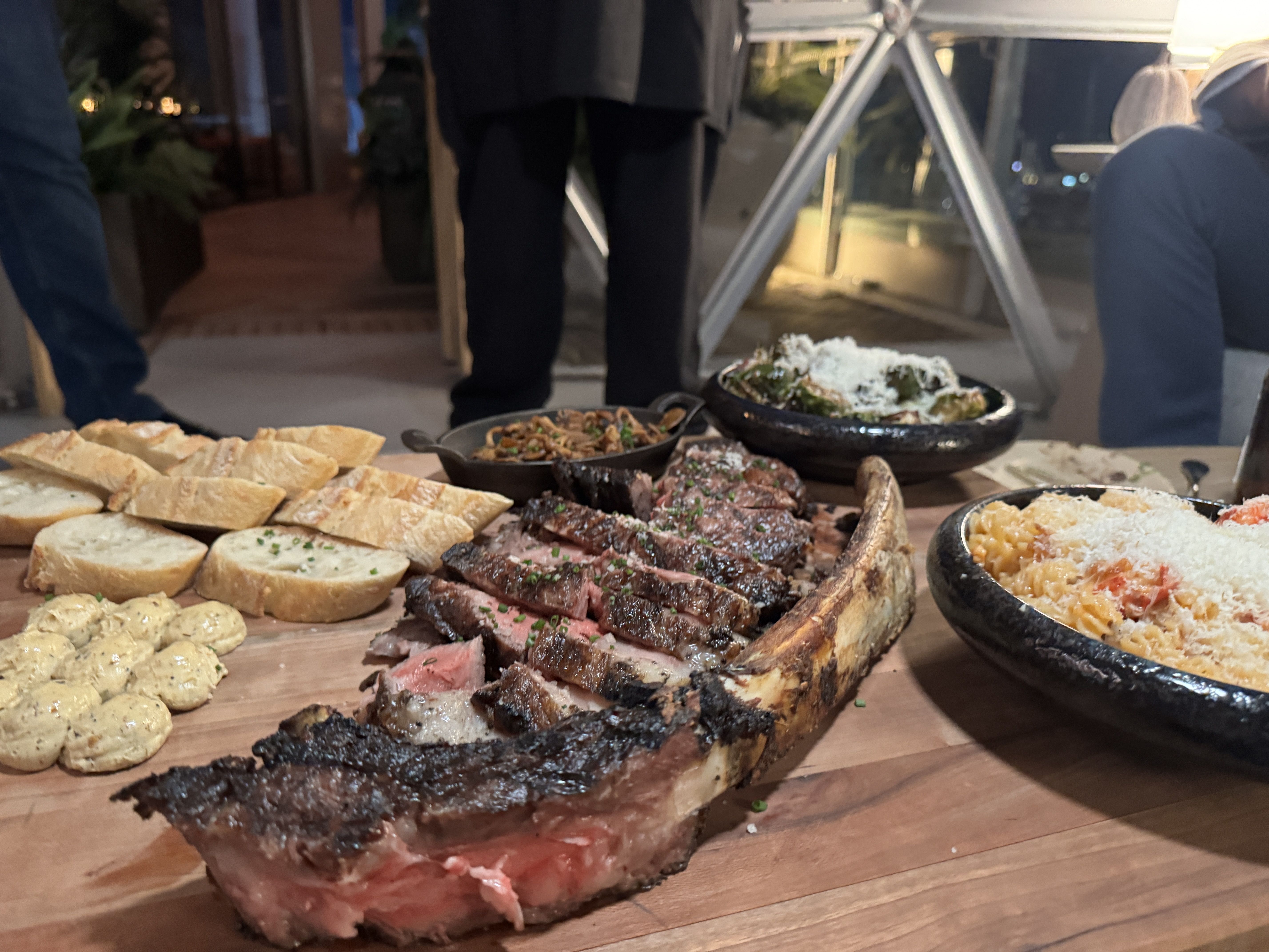 Sliced medium rare grilled steak with bread slices, mustard dollops, and bowls of pasta and vegetables on a wooden board, with people standing in the background.