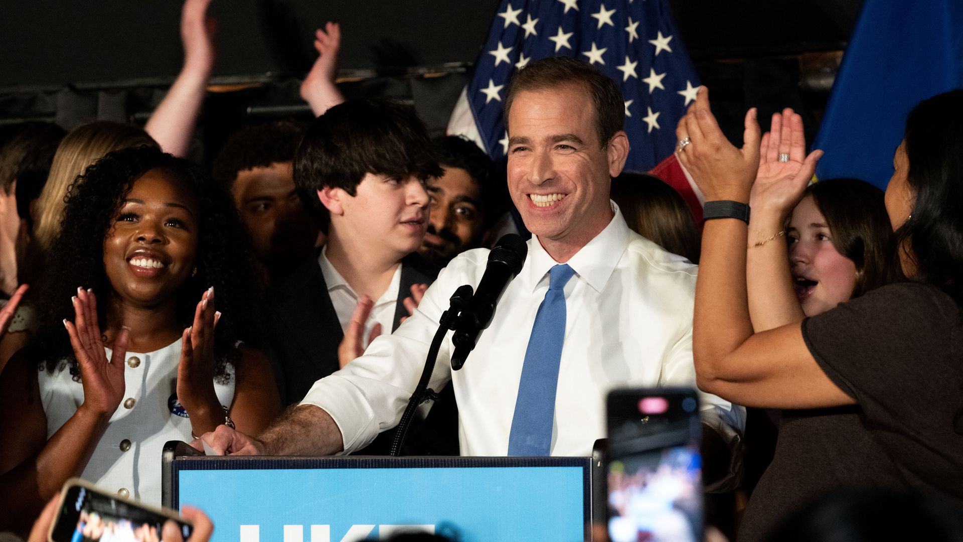 Smiling man in a white shirt and blue tie speaks at a podium with microphones, flanked by a cheering crowd. An American flag backdrop, raised hands, and people recording on phones.