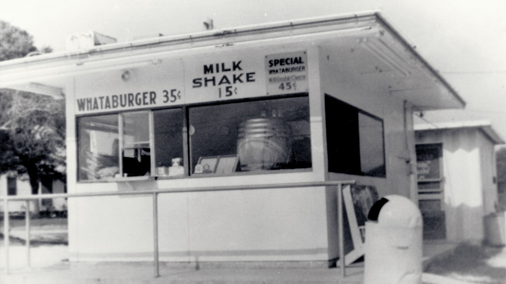 Black and white photo of an old Whataburger stand with prices: Whataburger 35¢, Milk Shake 15¢, and a special Whataburger with double cheese for 45¢. A trash can is in front.