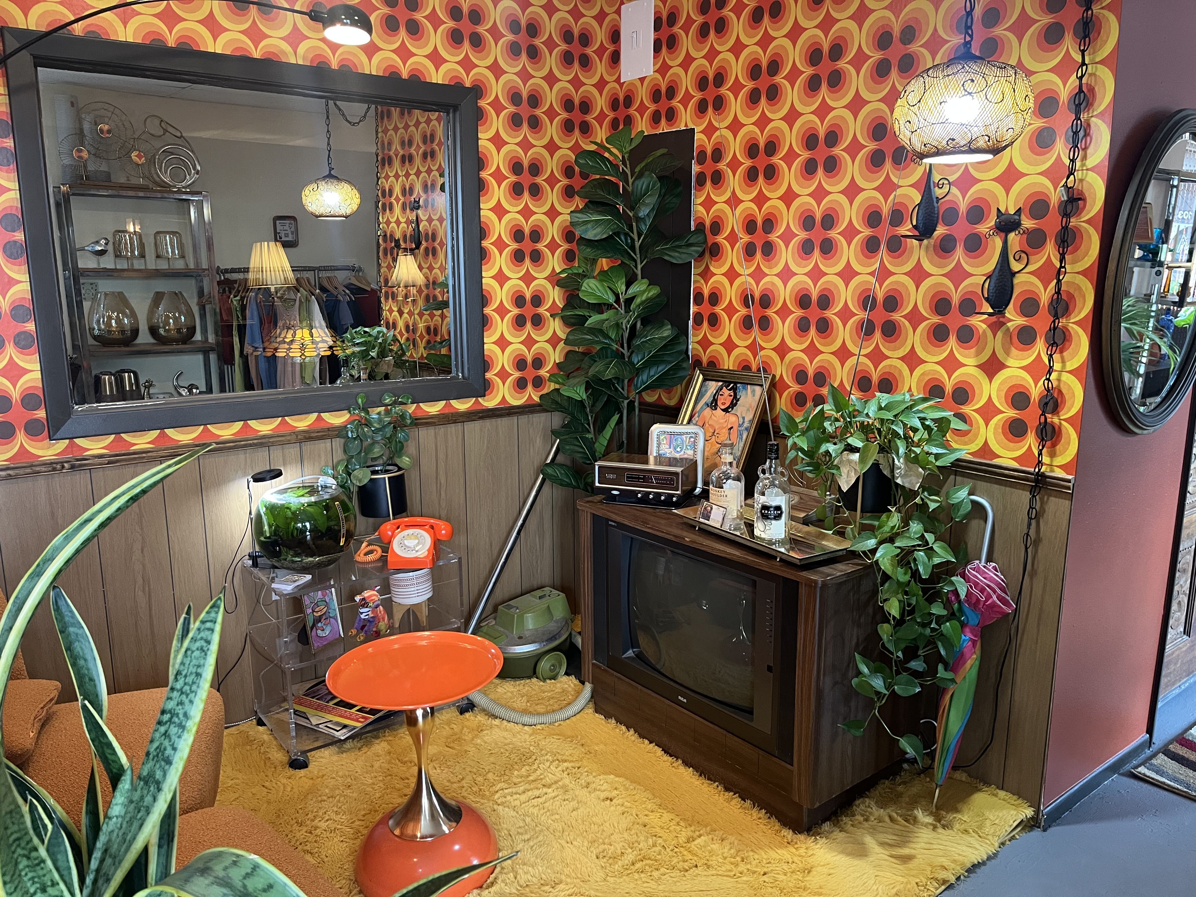 Bright retro interior with orange and yellow circular wallpaper, yellow shag rug, mid-century furniture, a hanging lamp, potted plants, and a mirror reflecting shelves of vintage decor.