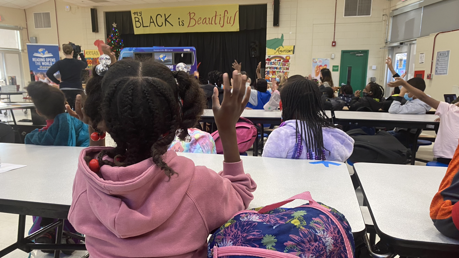 Young students sitting with their backs to the camera in a school cafeteria wave to a speaker getting ready to address them.