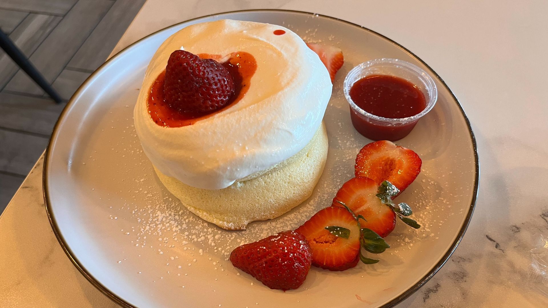A plate dessert featuring a small sponge cake topped with whipped cream and a whole strawberry, finished with strawberry sauce; sliced strawberries and a small cup of sauce decorate the plate.