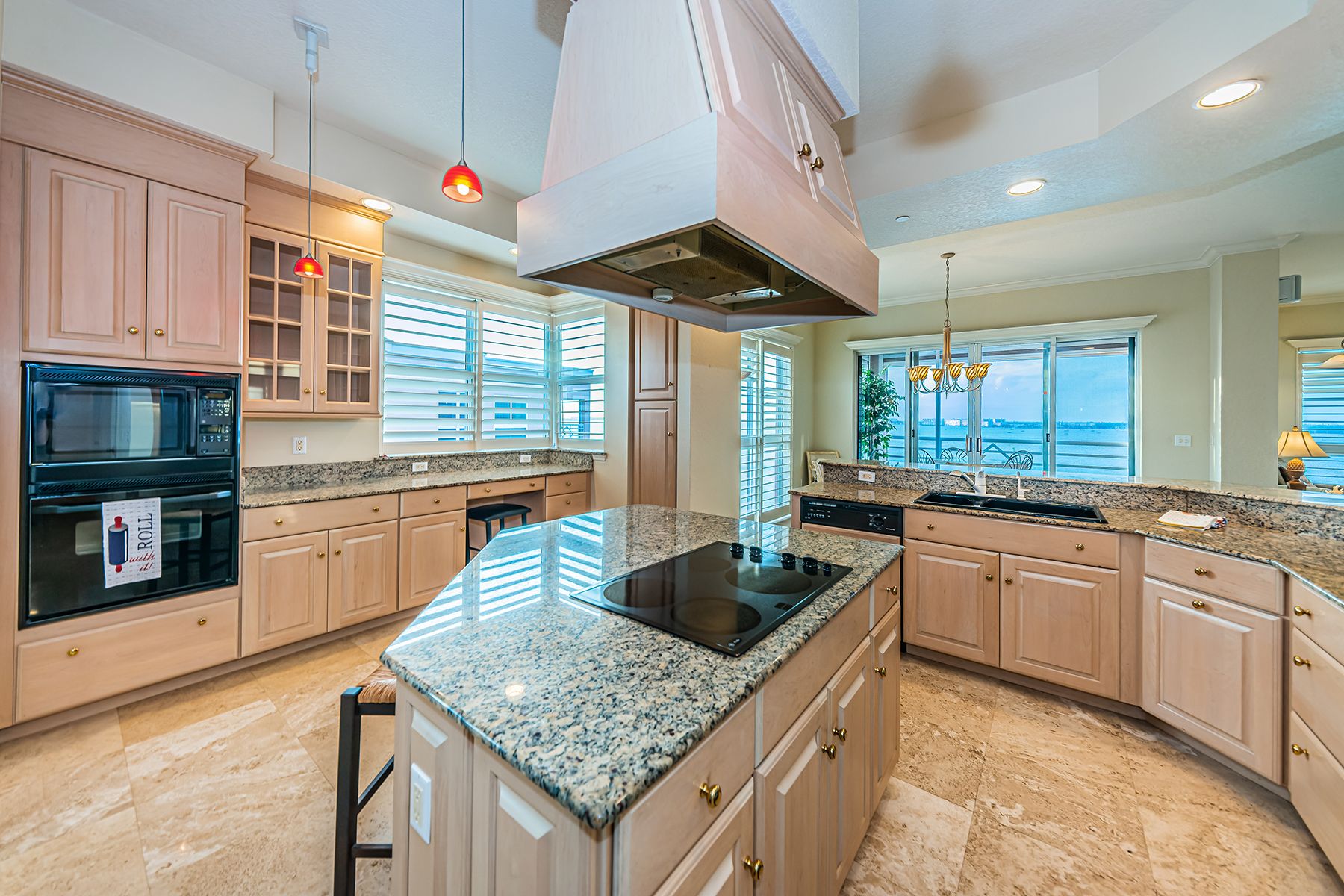 A kitchen with light wood cabinets and black-and-tan granite countertops with windows overlooking the water.