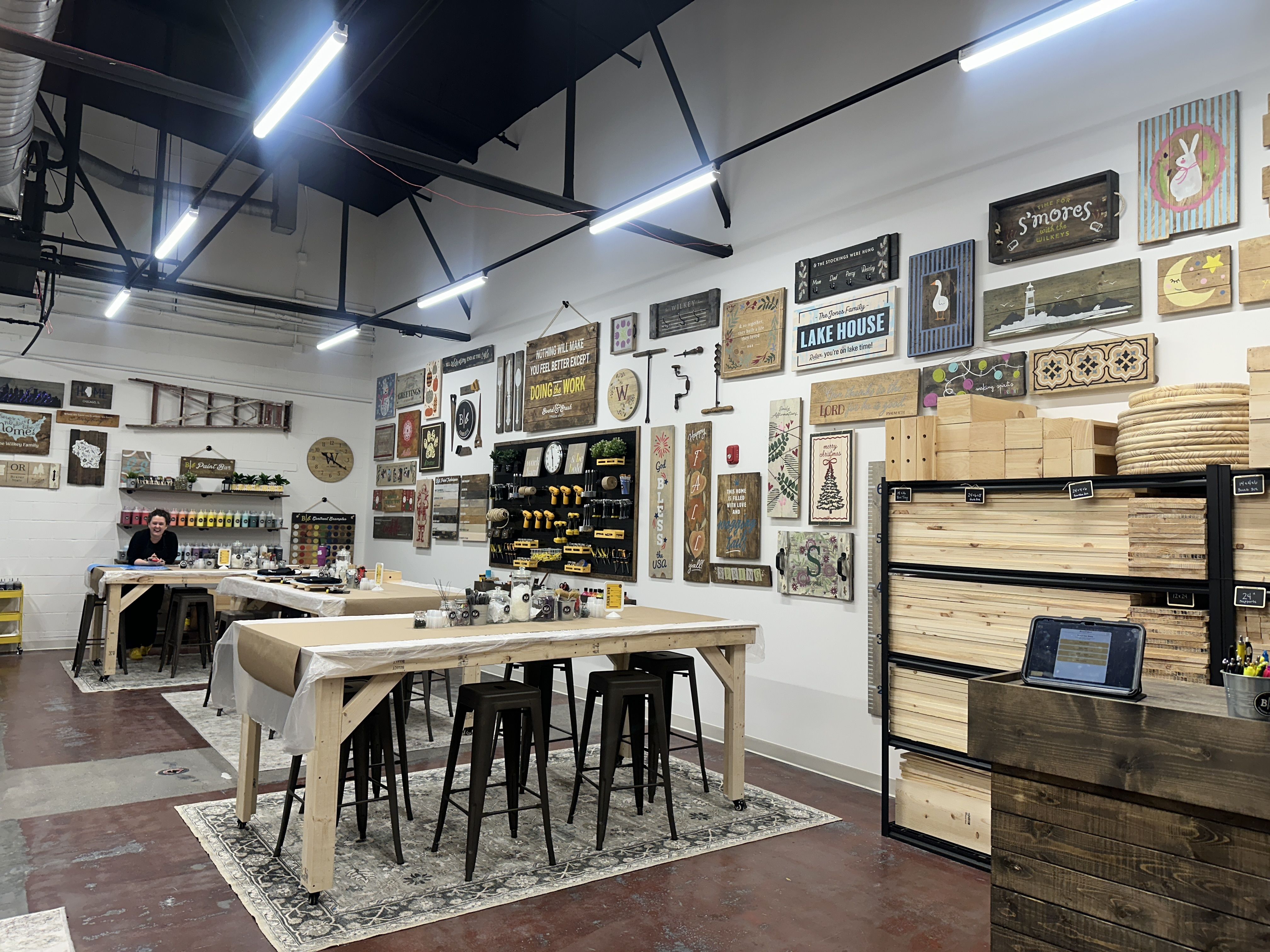 Board & brush. Bright woodworking shop with long tables and black stools. Walls display decorative wooden signs and art. A person works at a back table; right side shows stacked lumber on shelves.