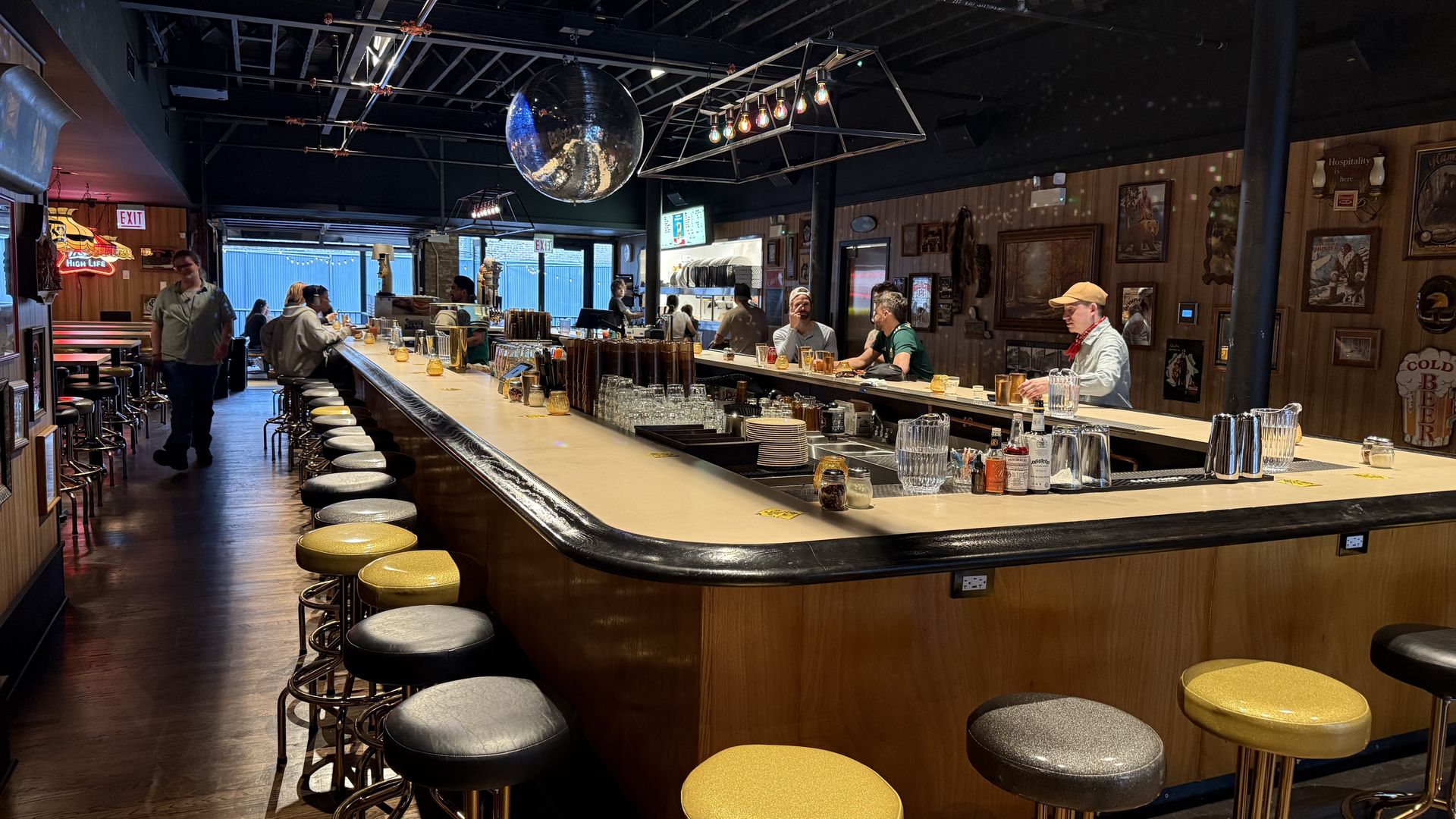 Retro-style bar with a long wooden counter and yellow, gray, and black stools. Patrons sit at the bar; a disco ball hangs from the ceiling. Framed pictures cover the wood-paneled wall.