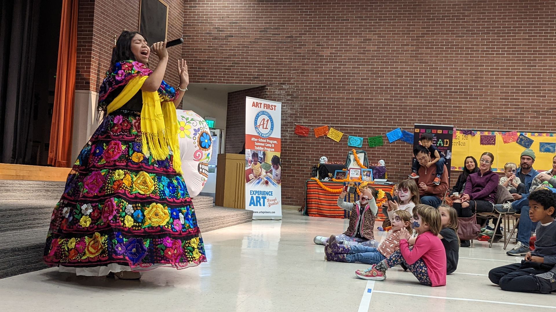 A girl in a colorful dress with a hoop skirt sings in front of children in an elementary school gymnasium.