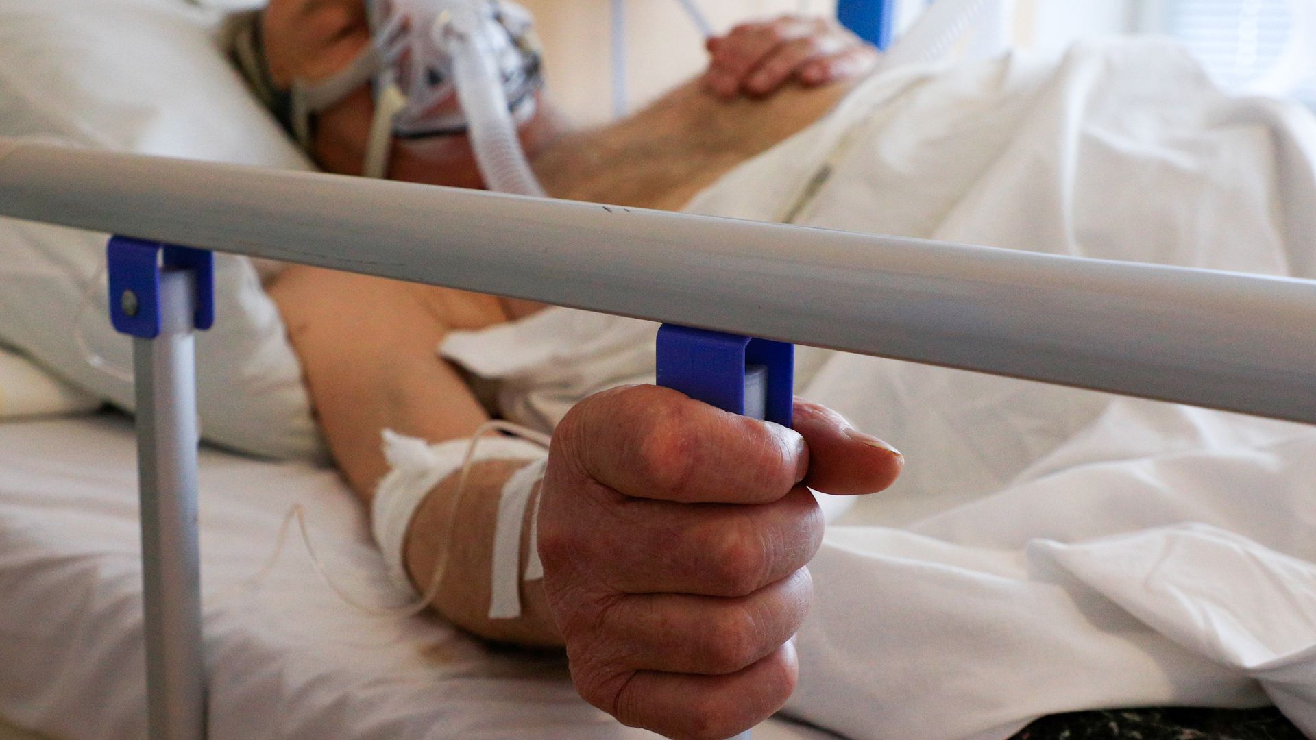 Photo of a patient wearing a ventilator, lying in a hospital bed and holding a blue device in their right hand