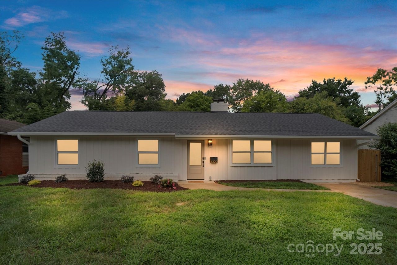 Single-story white house with dark gray roof at sunset, illuminated windows, a front door with a light, small garden plants, green lawn, and trees in the background.