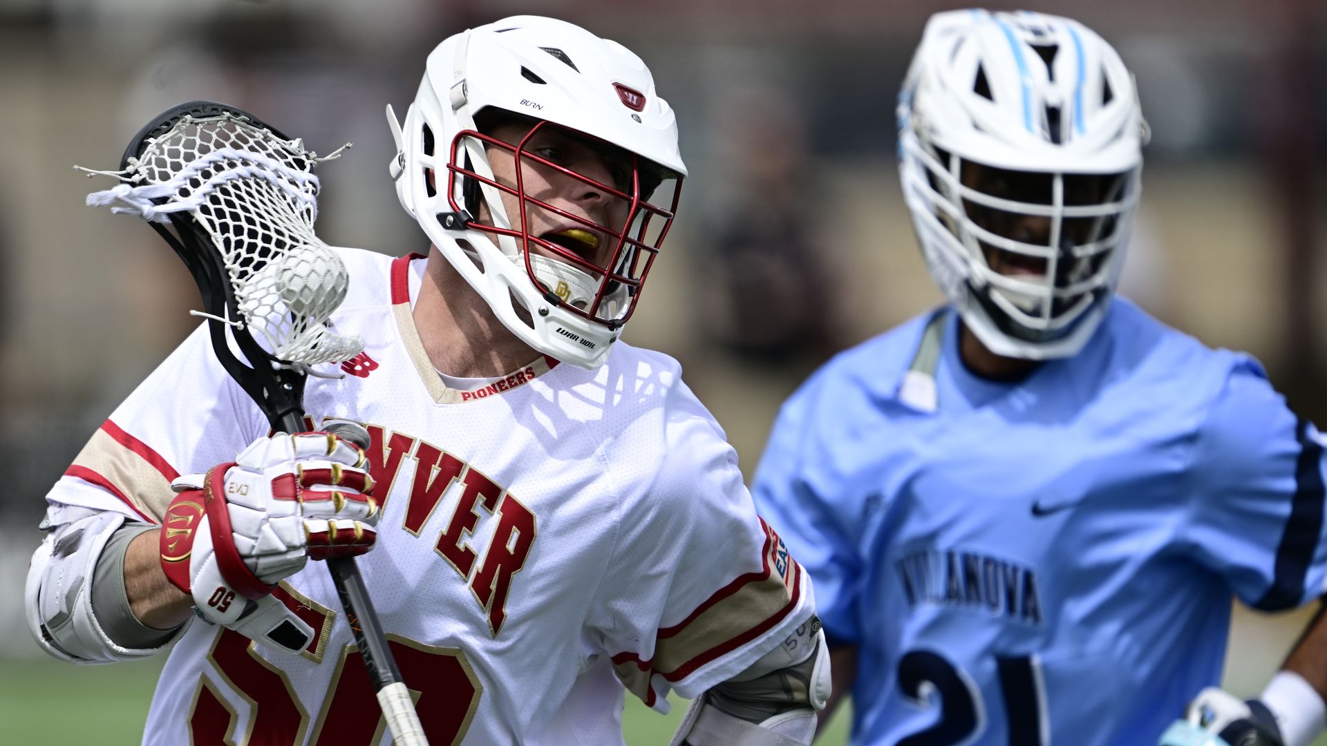 A man in a helmet and a lacrosse stick runs in front of another man in a helmet. 