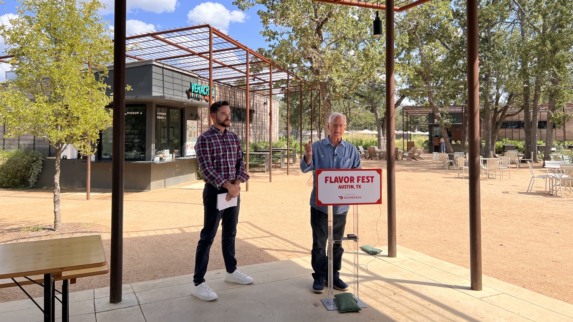Outdoor scene under a brown metal pergola; two men stand near a sign reading Flavor Fest Austin, TX, with a building, trees and blue sky in the background.