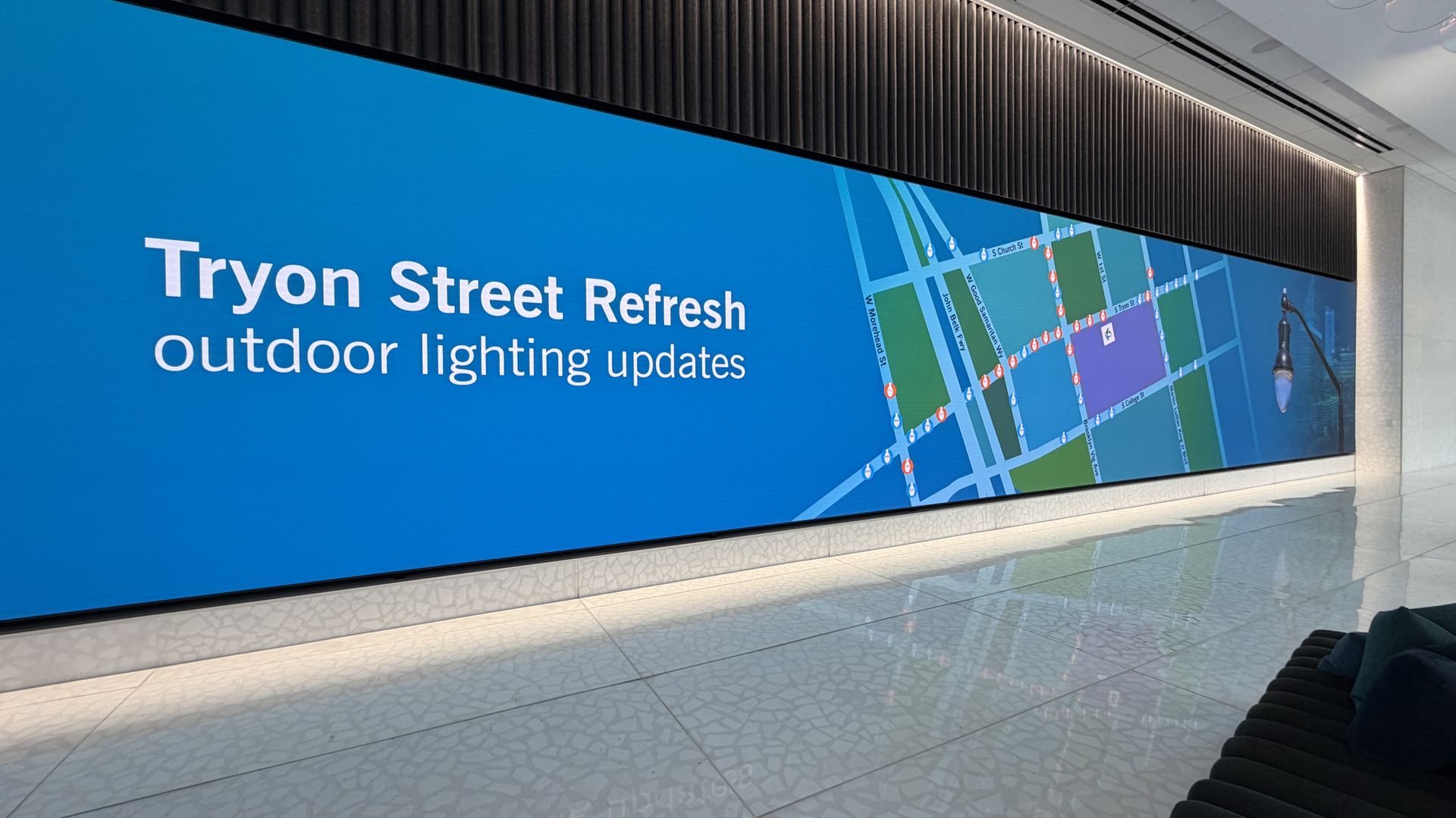 Indoor digital display with a blue background showing "Tryon Street Refresh outdoor lighting updates" and a map with marked lighting locations in red and blue icons.